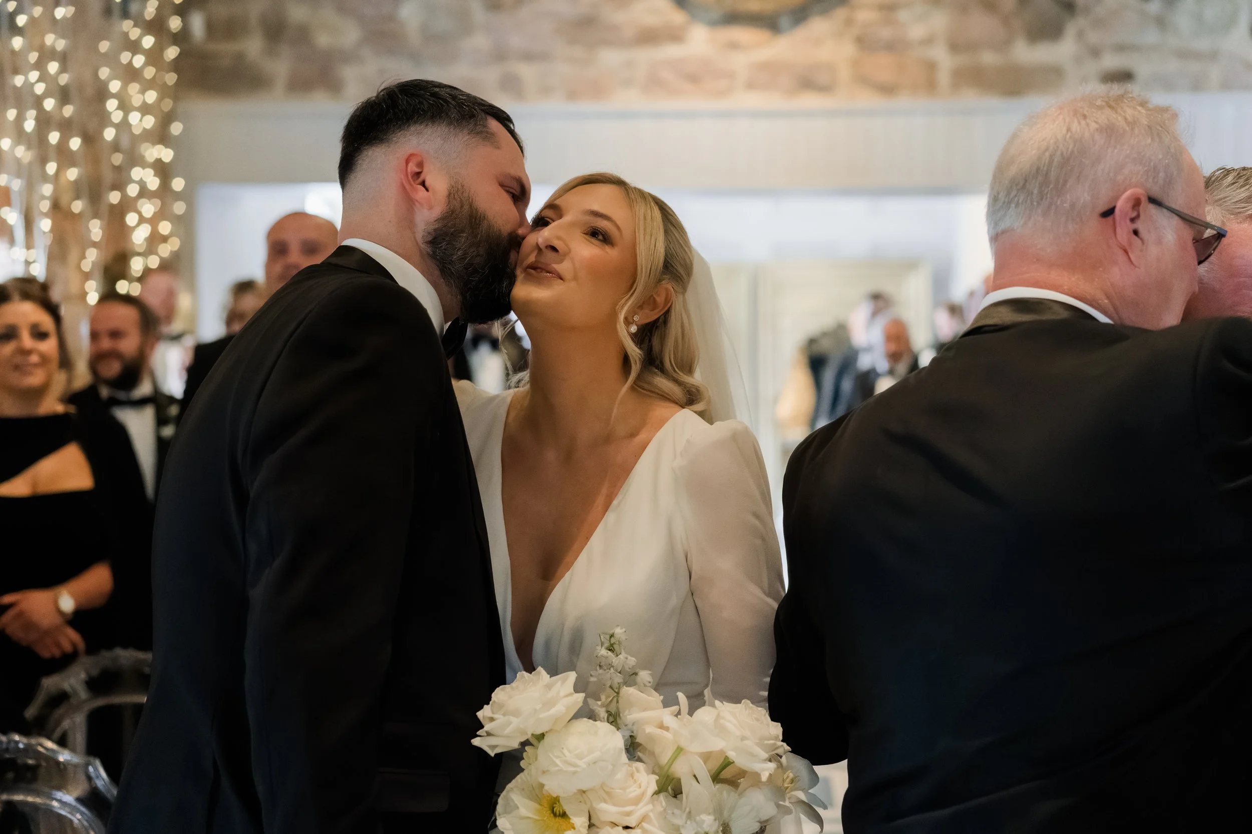 A bride and groom share a kiss at their wedding reception, surrounded by guests in formal attire.