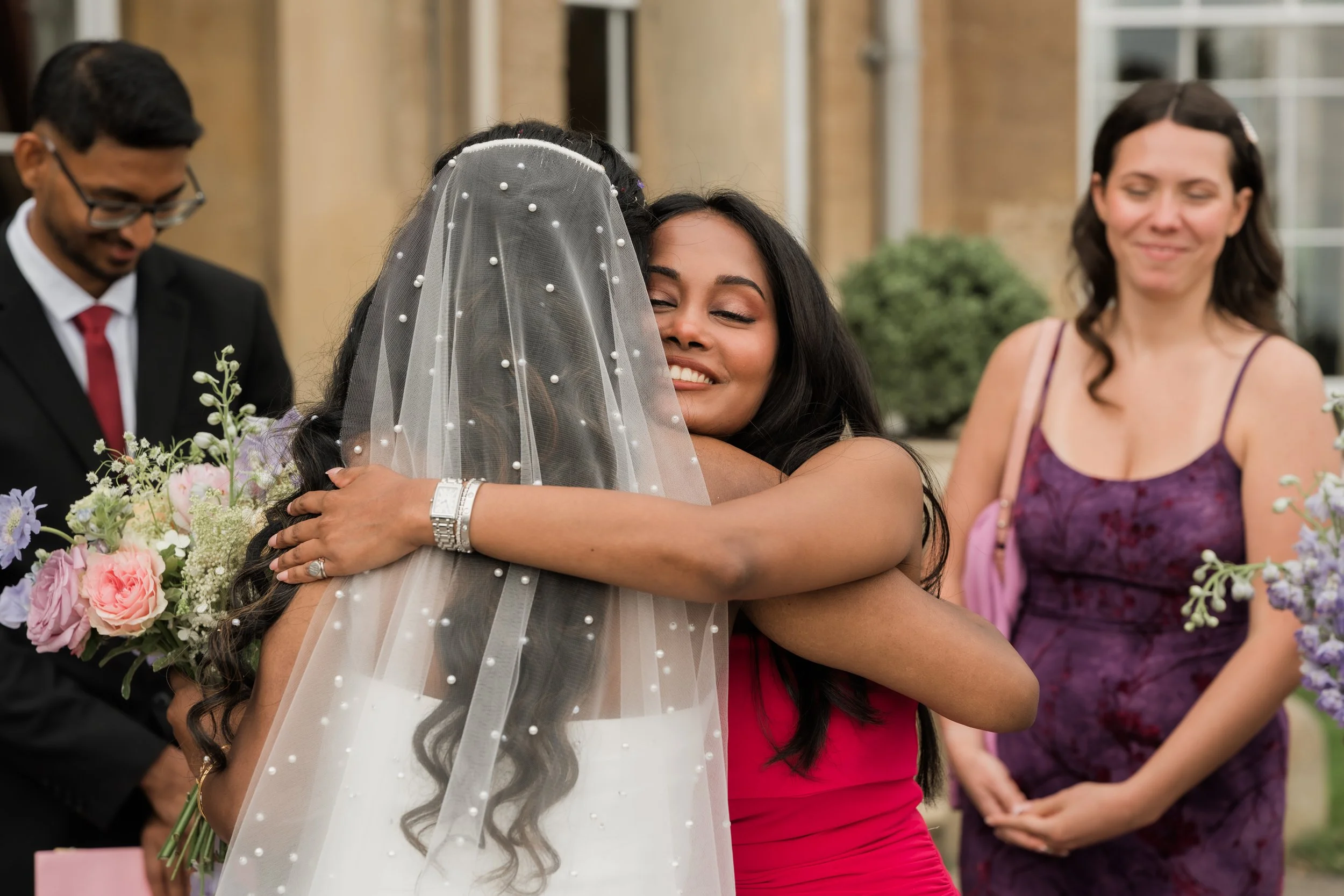 Two women hugging at a wedding, one in a wedding dress and veil, the other in a red dress, with guests and flowers in the background.