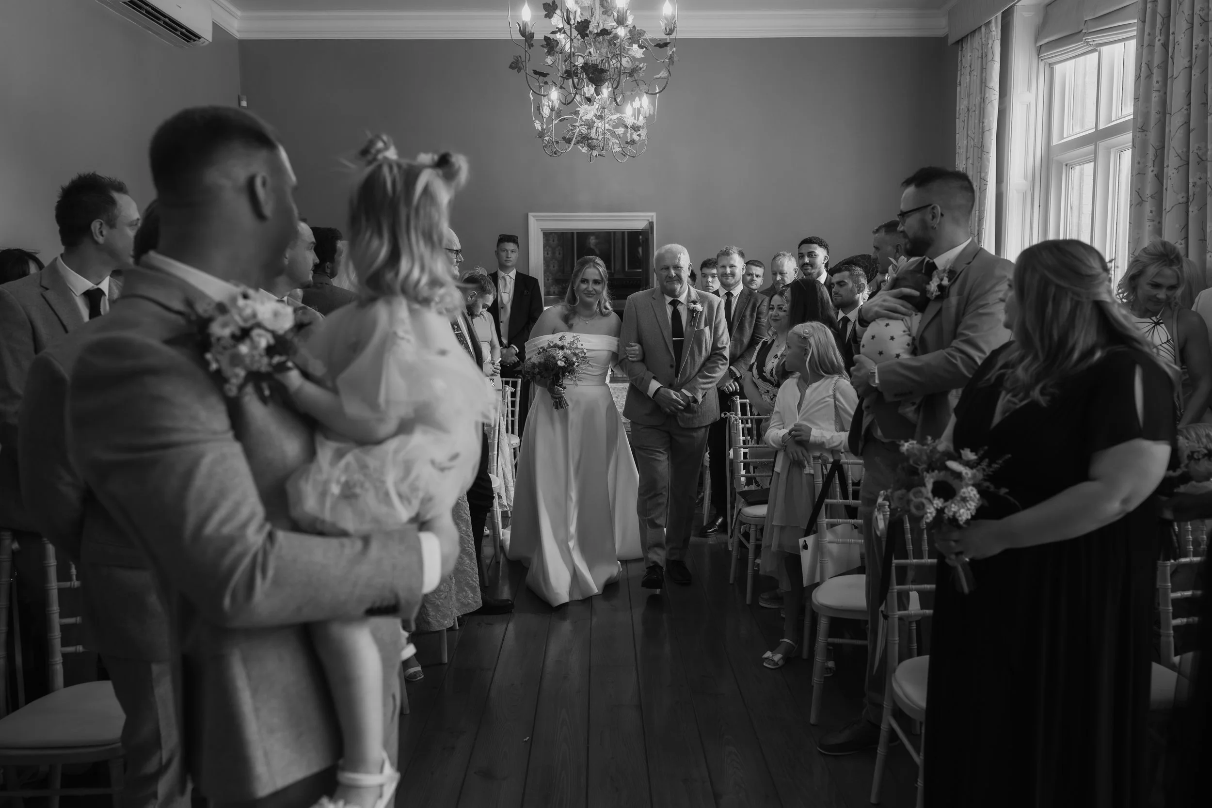 A black and white photo of a wedding ceremony with guests standing in a room. The bride is walking down the aisle holding a bouquet, accompanied by a man. Guests are watching, some holding flowers, in a room with a chandelier and large windows.
