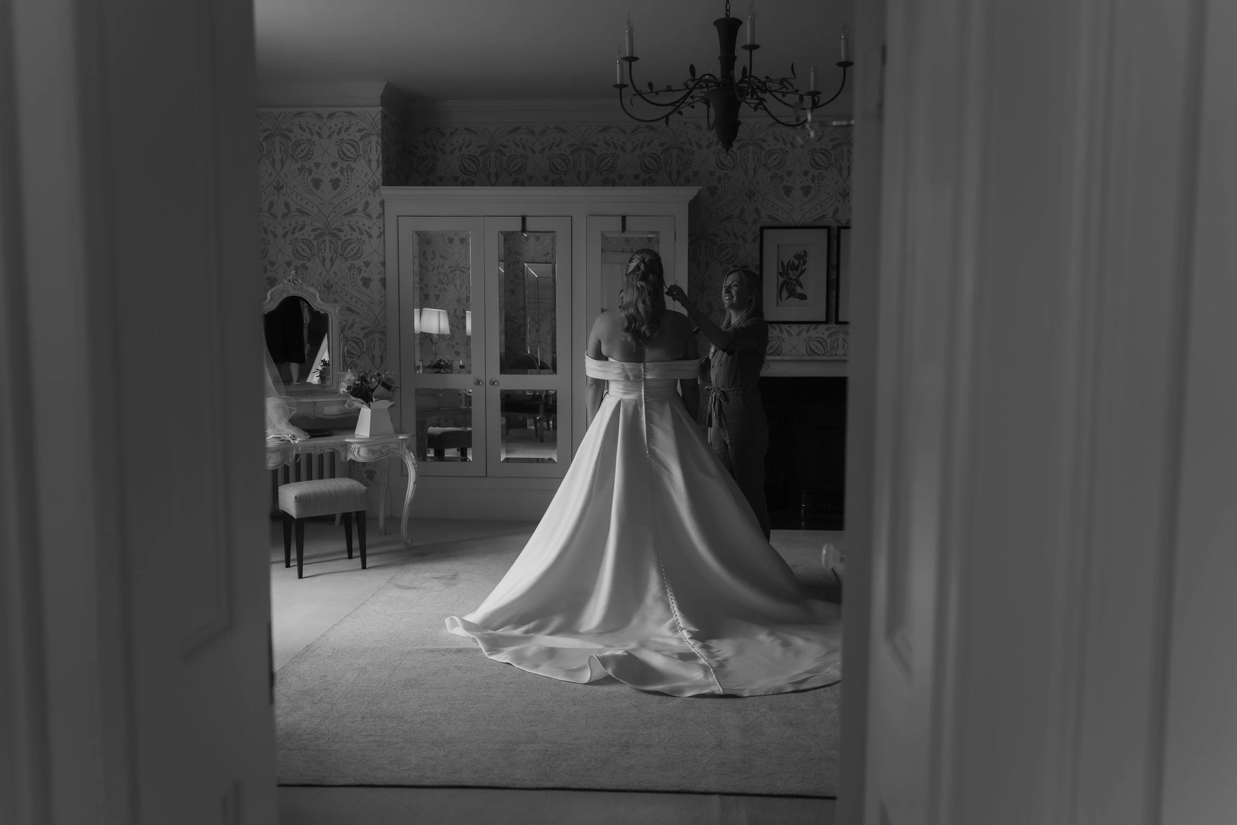 A bride in a wedding dress standing in front of a mirror with a woman adjusting her makeup.