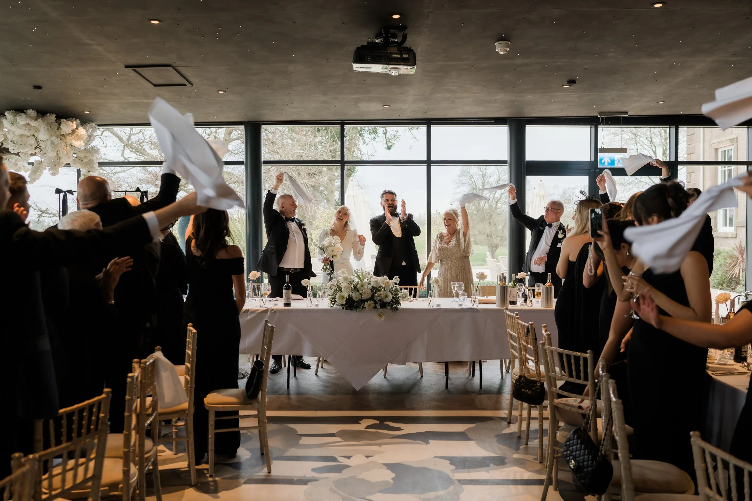 People celebrating at a wedding reception in a modern indoor venue with large windows, a long reception table, and a floral centerpiece, as guests throw napkins in the air.