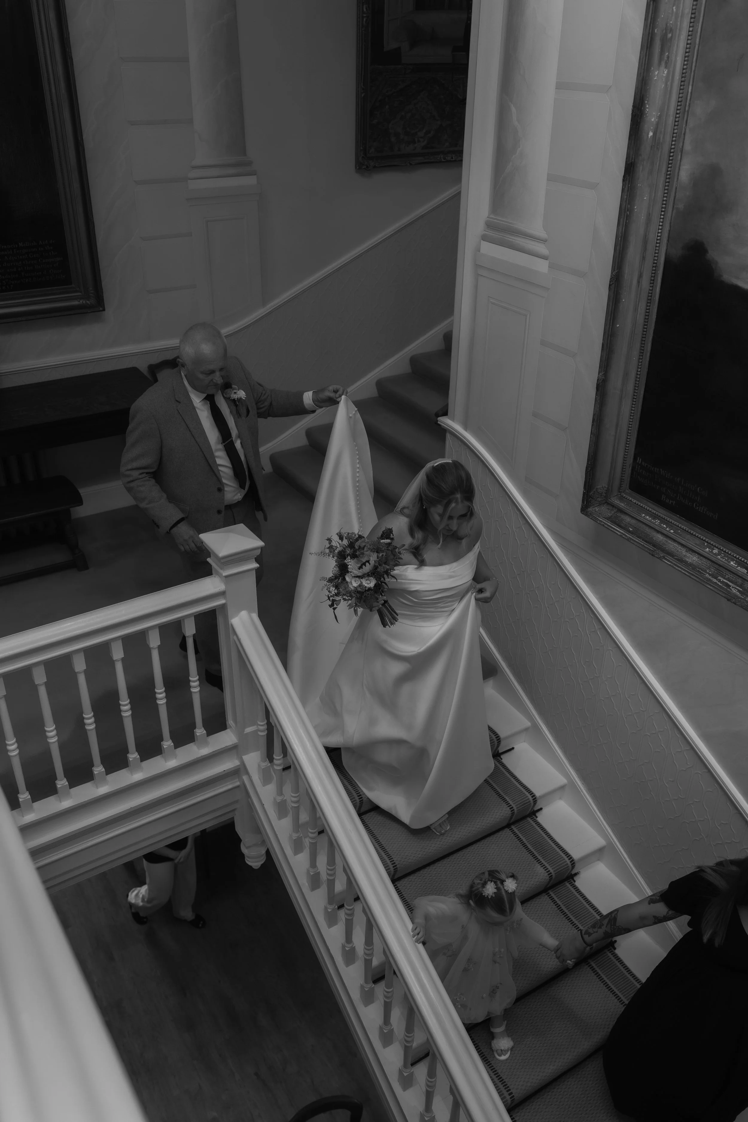 A bride in a wedding dress holding a bouquet, ascending a staircase while someone holds a flag behind her, with a young girl reaching up to hold her hand, in an elegant indoor setting.