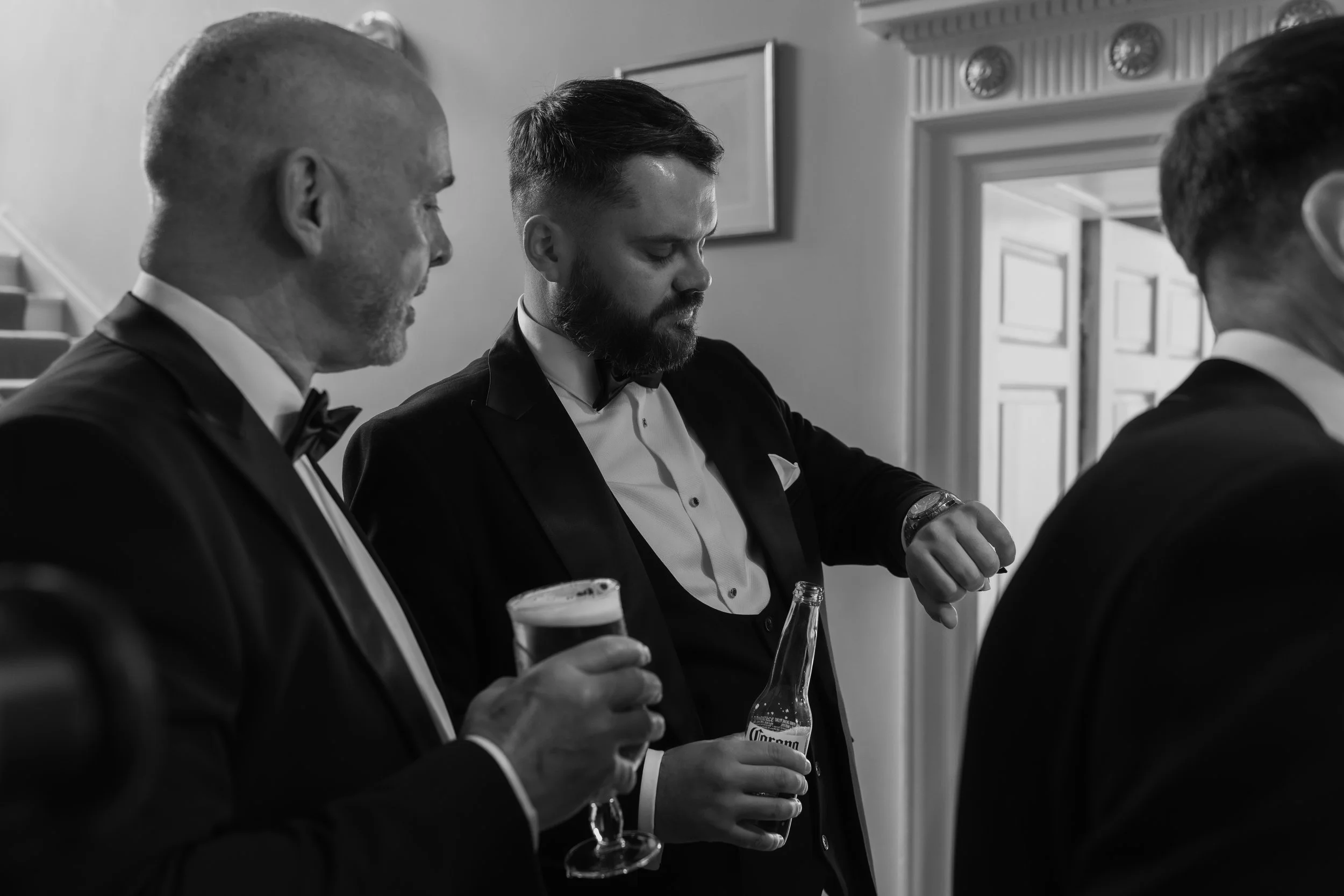 Three men in tuxedos gathered indoors, one man looking at his watch, others holding drinks, suggesting a formal event or celebration.
