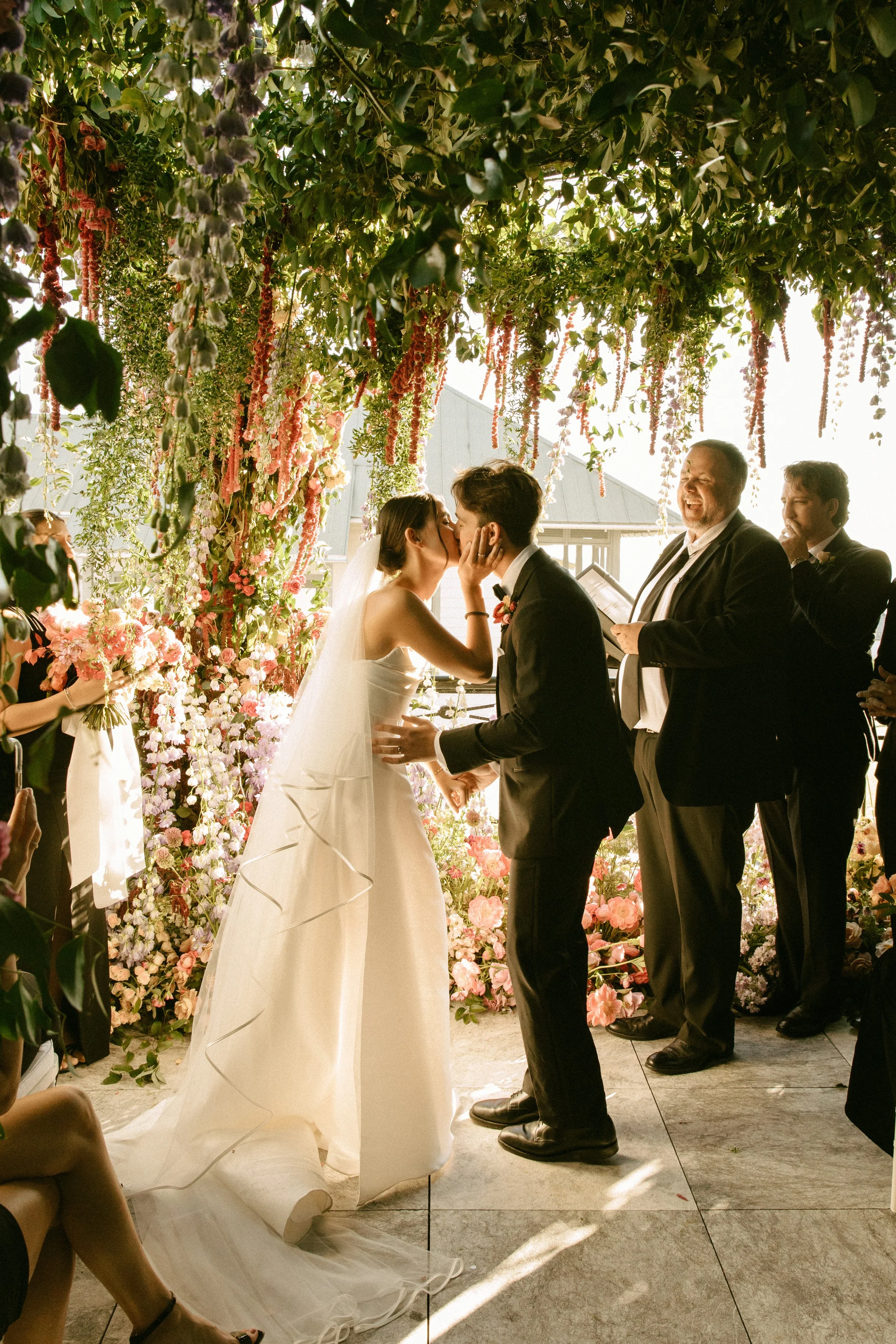 A wedding ceremony with a bride and groom kissing under a floral arch with pink and white flowers, surrounded by guests.