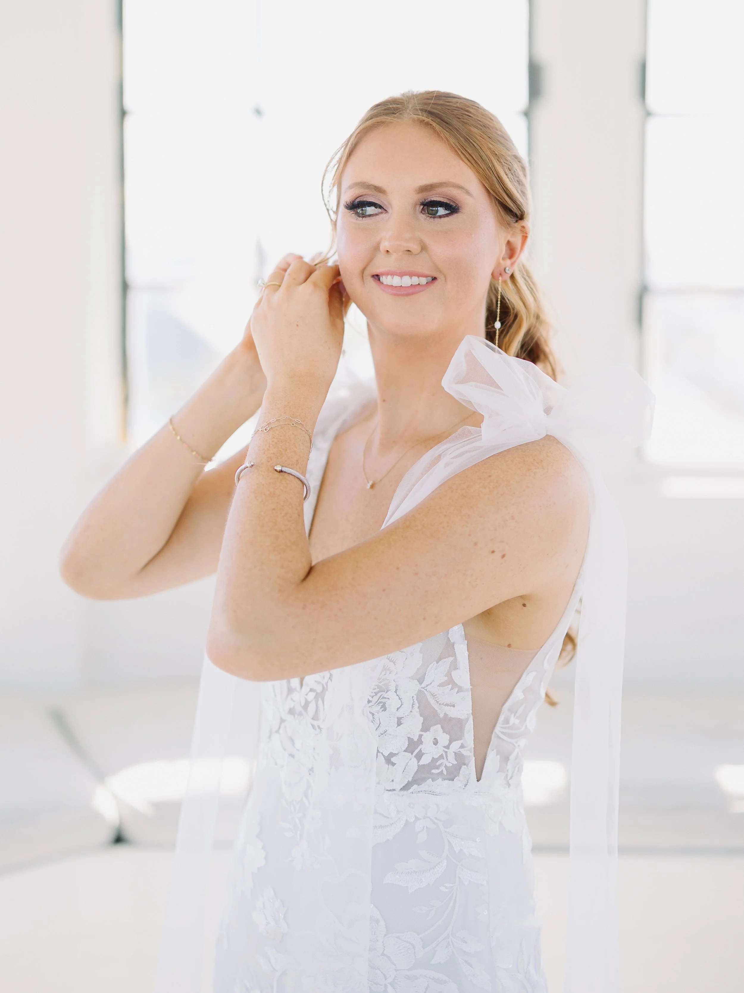 A woman in a white wedding dress with floral lace details, adjusting her earring indoors with large windows in the background.
