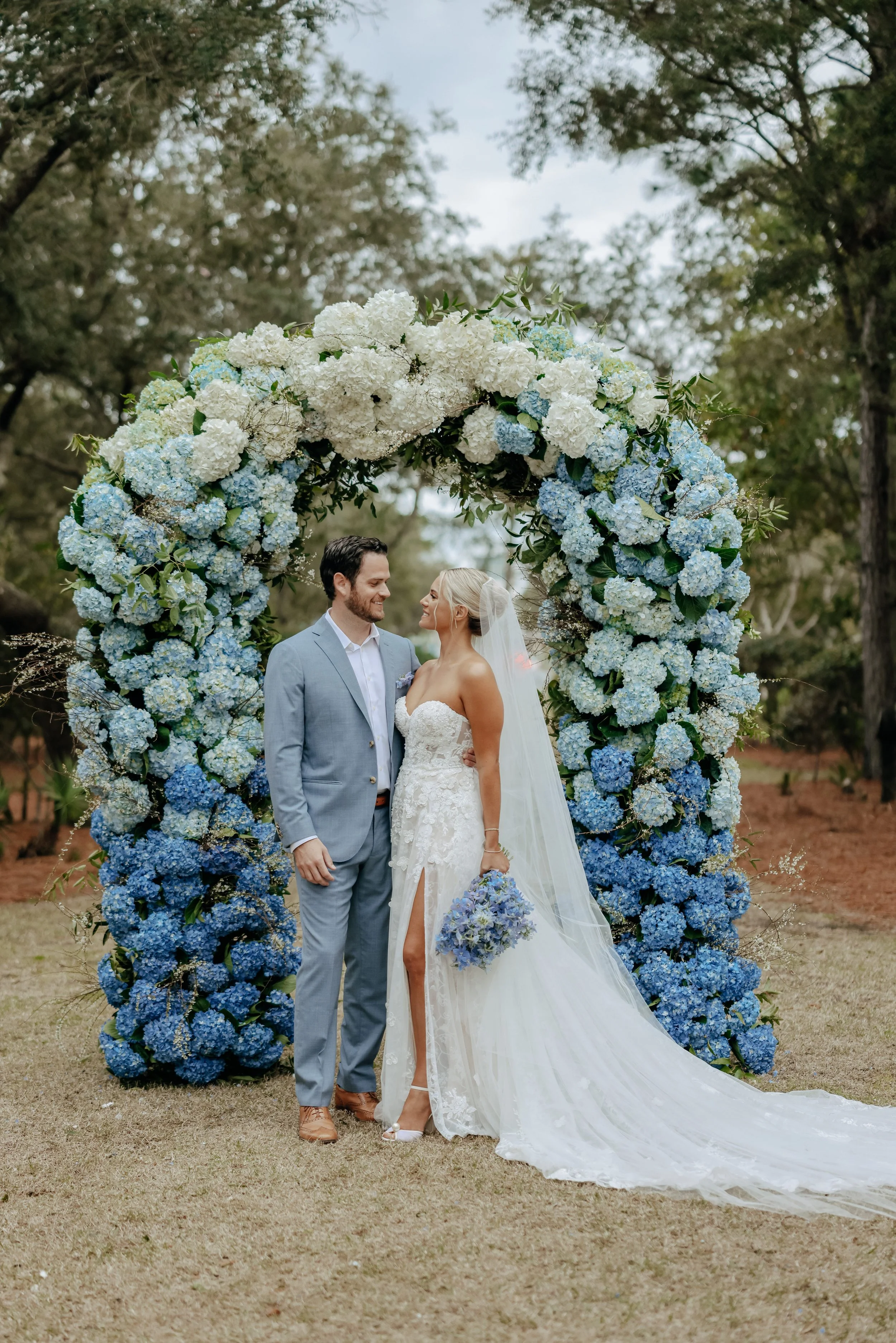 A bride and groom holding hands and smiling at each other standing beneath a large arch made of white and blue hydrangeas in a garden.