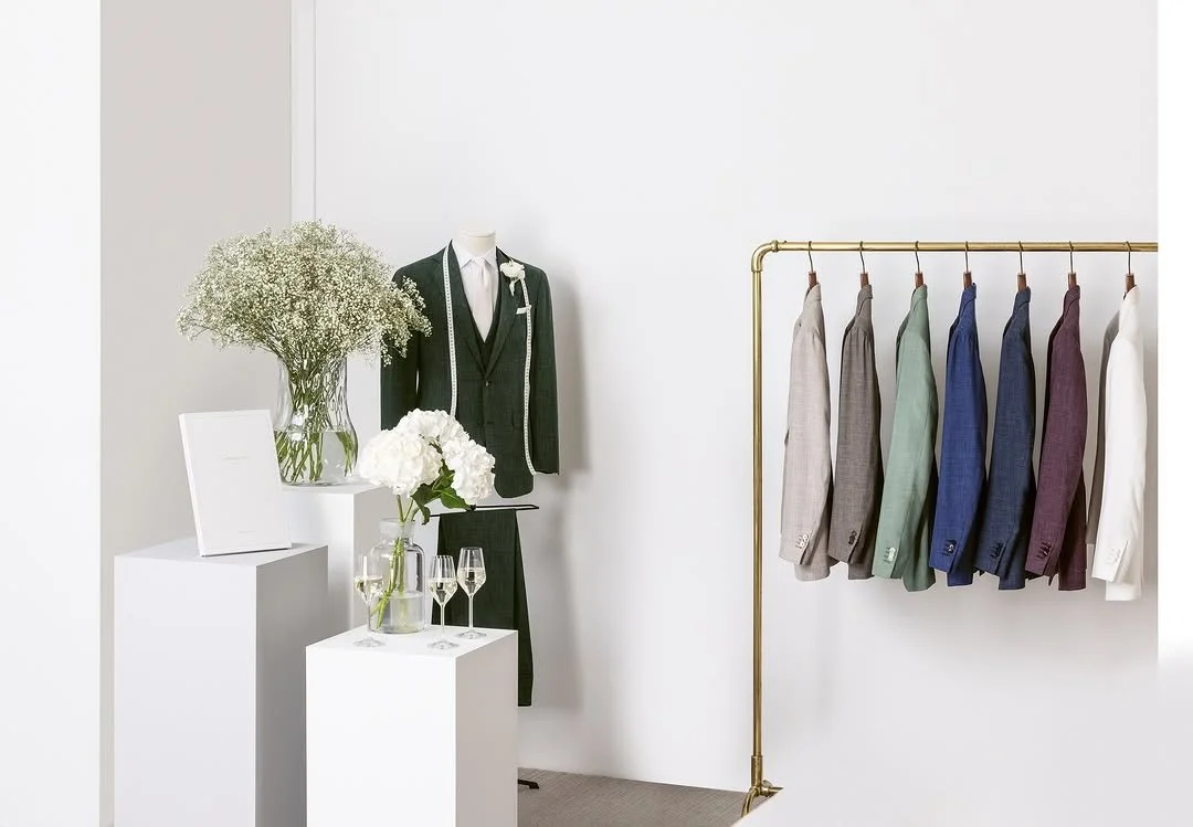 Display of men's suits and tuxedos with colorful blazer jackets hanging on a gold clothing rack, a mannequin dressed in a black tuxedo with a measuring tape, and white vases with flowers and glasses on white pedestals.