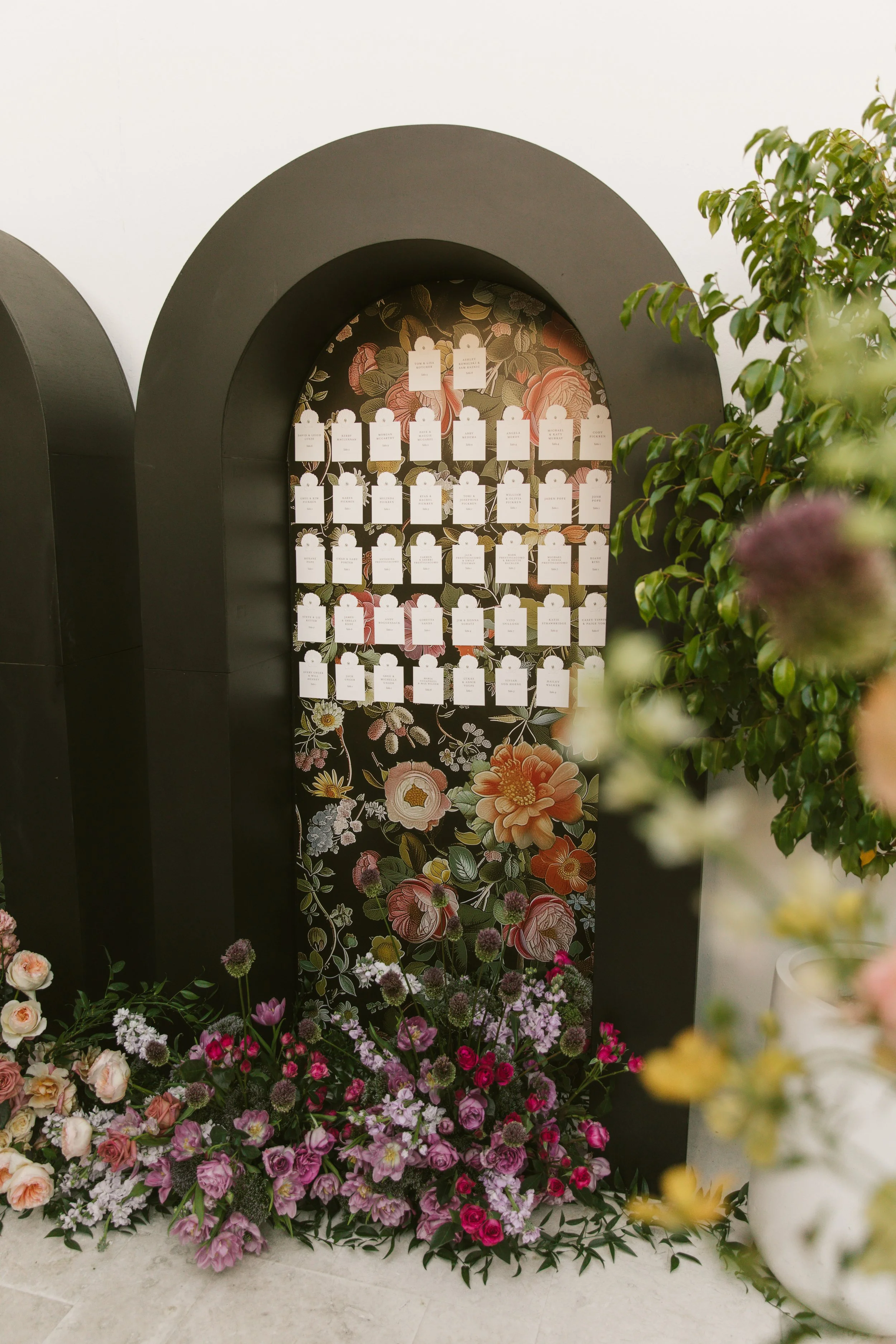 A display board with floral wallpaper, holding several white tags, surrounded by a large arrangement of pink and purple flowers and green plants.