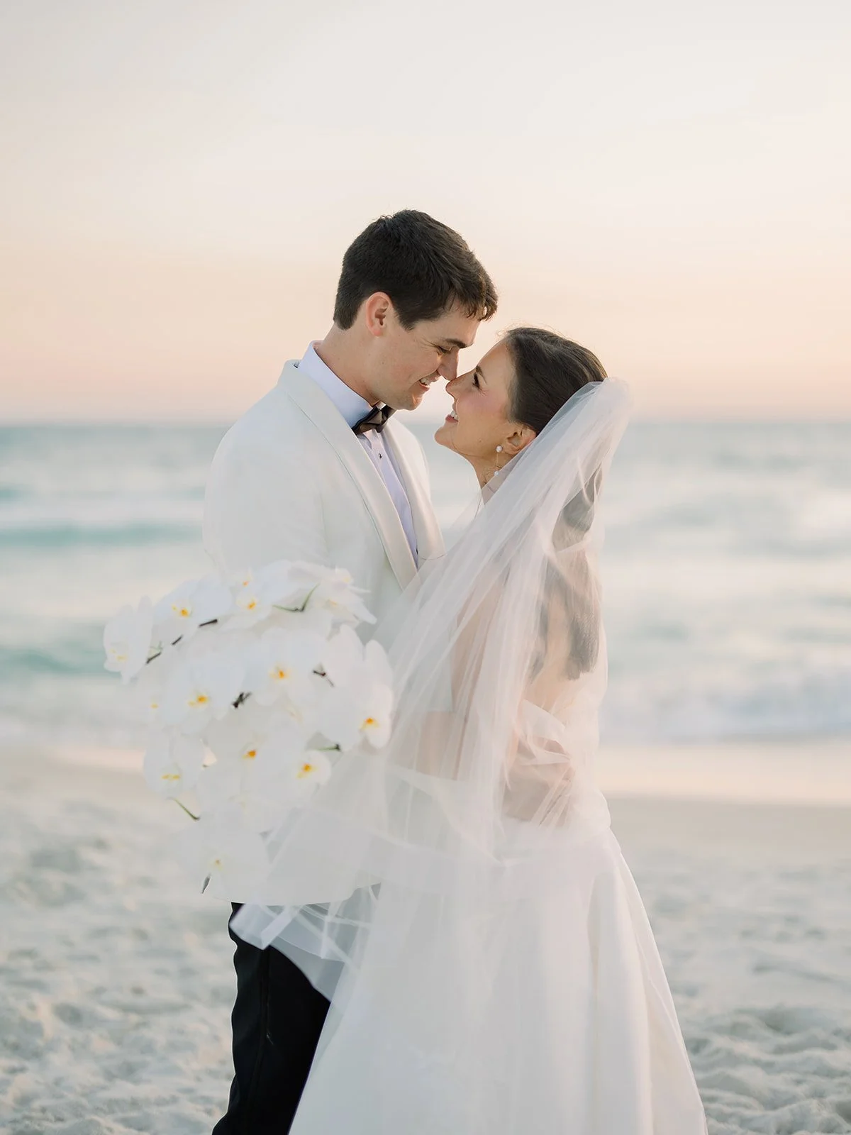 A newlywed couple in wedding attire on the beach, smiling and about to kiss with ocean waves and a pink sunset in the background.
