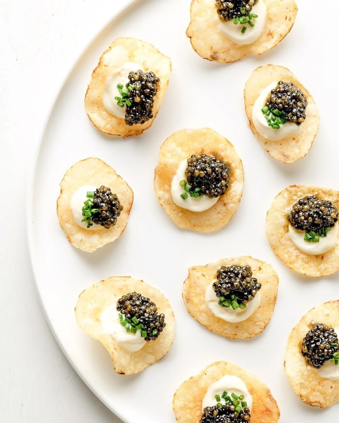Canapés with crispy potato chips topped with sour cream, caviar, and chopped chives on a white plate.