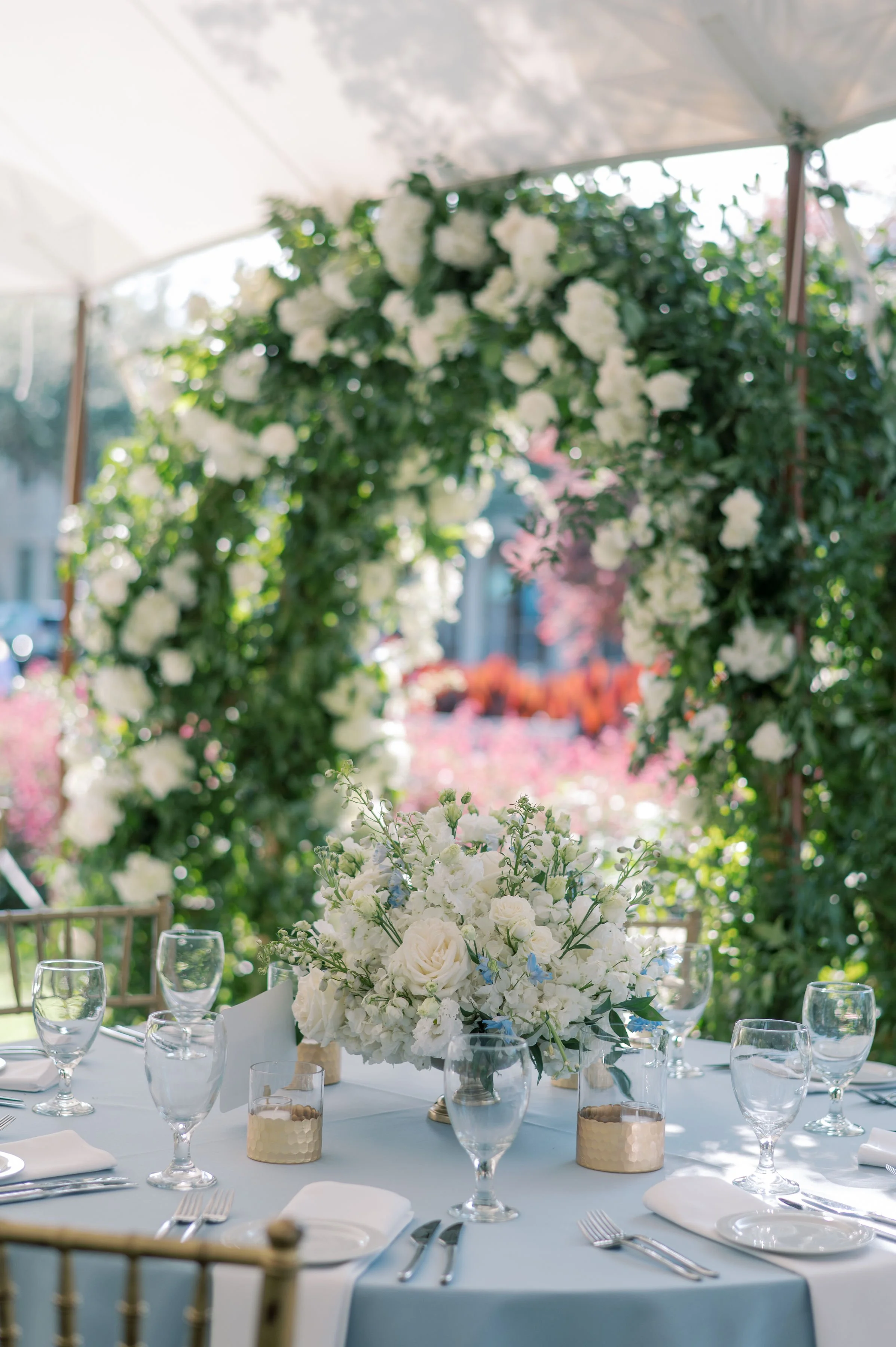 Elegant outdoor table setup with white floral centerpiece, glassware, plates, and cutlery under a white canopy, decorated with a white flower arch, for a wedding or special event.