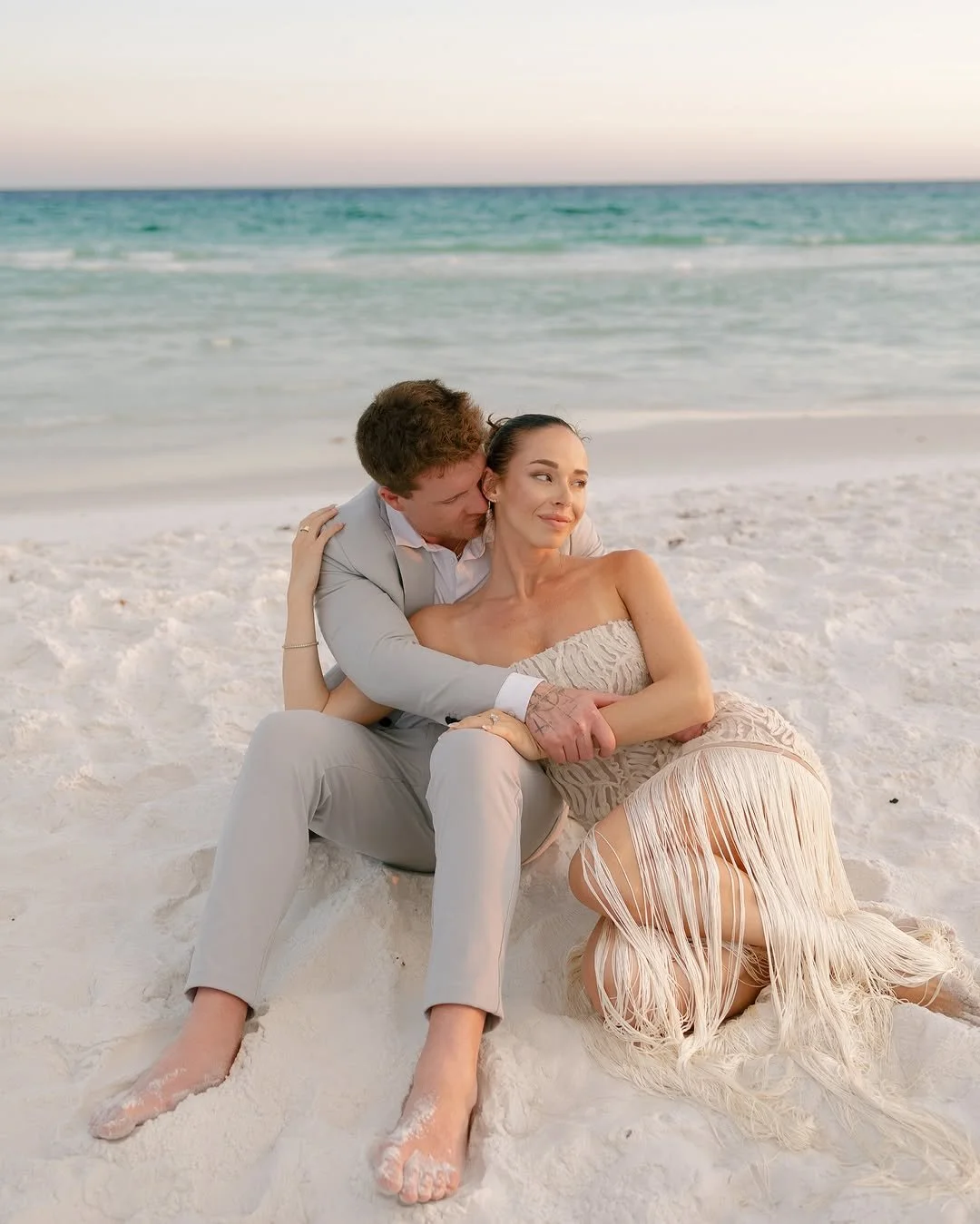 A couple sitting on white sand at the beach, with the ocean and sky in the background. The man in a light grey suit is kissing the woman's cheek, who is wearing a strapless, cream-colored dress with fringe detailing. They are barefoot, and the woman is sitting on the sand while the man is leaning in toward her.
