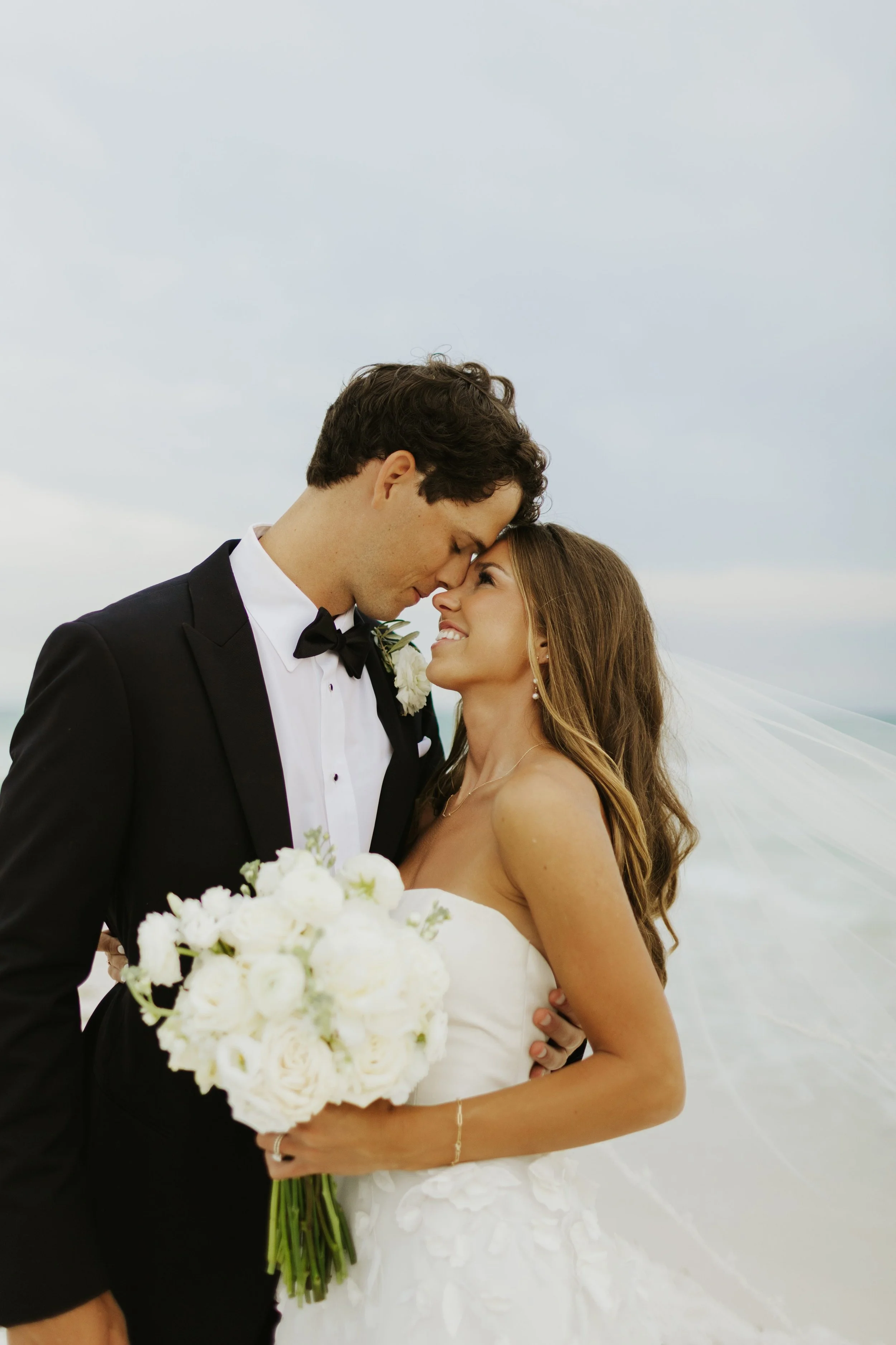A newlywed couple on their wedding day, standing close with foreheads touching, outdoors during the daytime, with the bride holding a bouquet of white flowers and the bride wearing a strapless white wedding gown and veil.