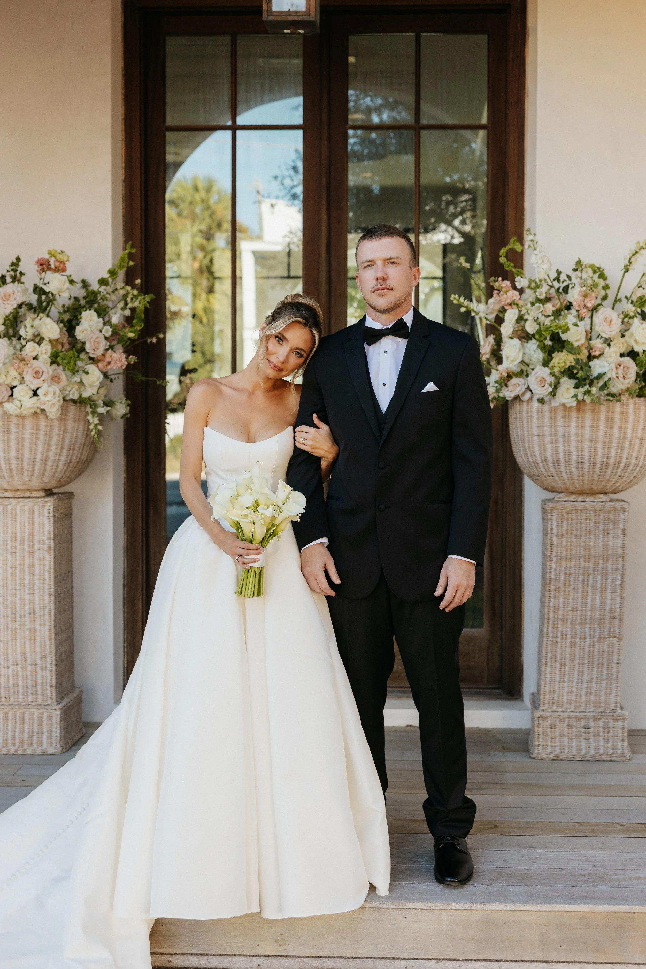 Bride and groom standing together at their wedding, with the bride holding a bouquet of white flowers and the groom in a black tuxedo, outside in front of a glass door with floral arrangements on either side.