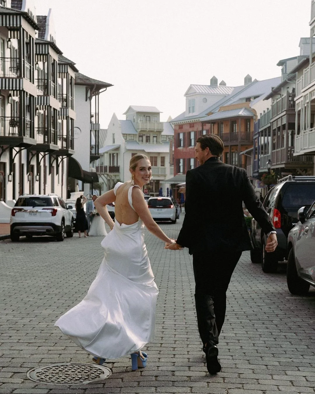 A bride in a white wedding dress and a groom in a black suit holding hands and walking on a cobblestone street with parked cars and colorful buildings in the background.