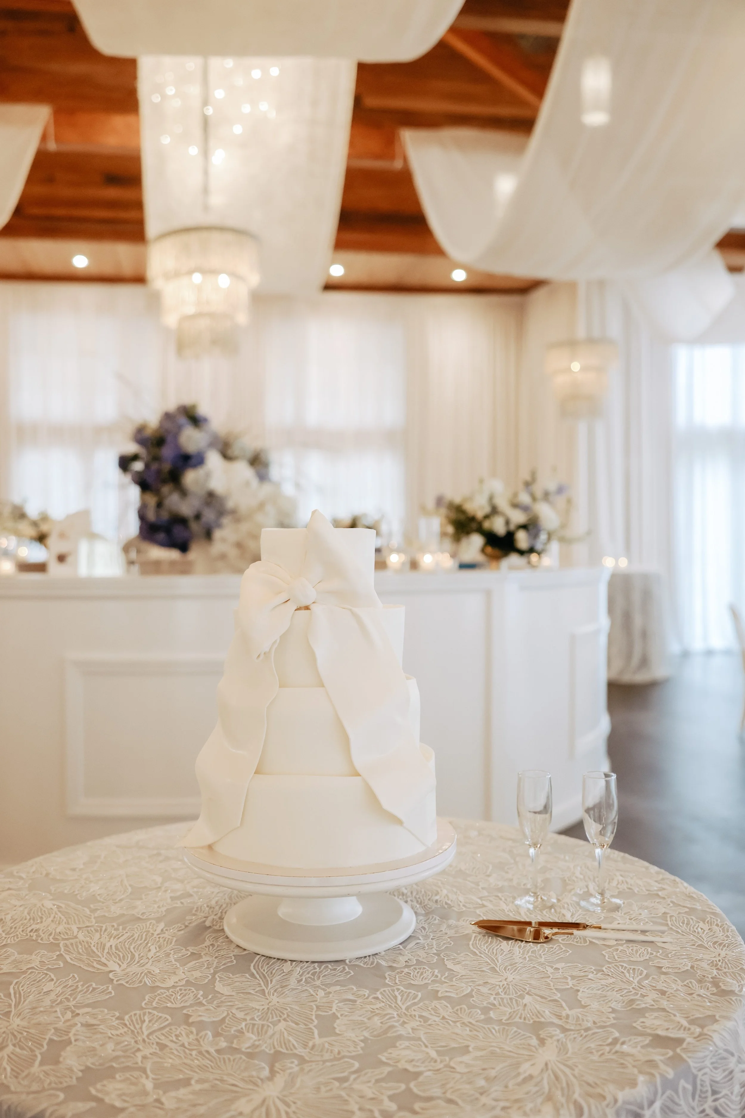 Three-tier white wedding cake with a large white bow on top, placed on a table with a lace tablecloth, in an elegant decorated reception hall.
