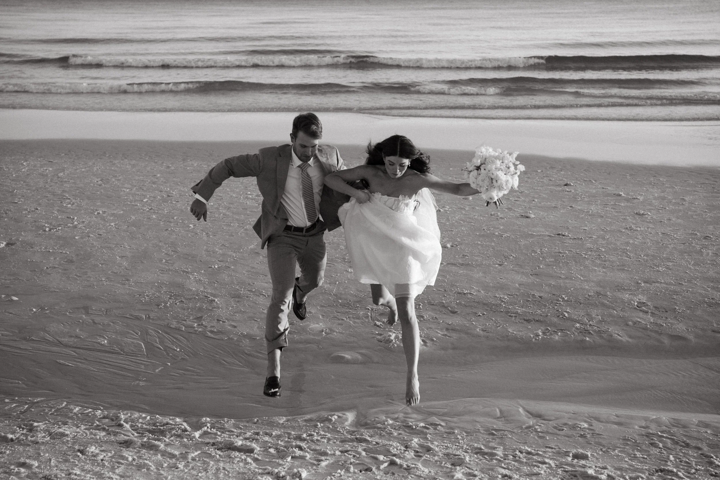 A black and white photograph of a newlywed couple running on the beach, with the bride holding a bouquet of flowers and the groom in a suit.