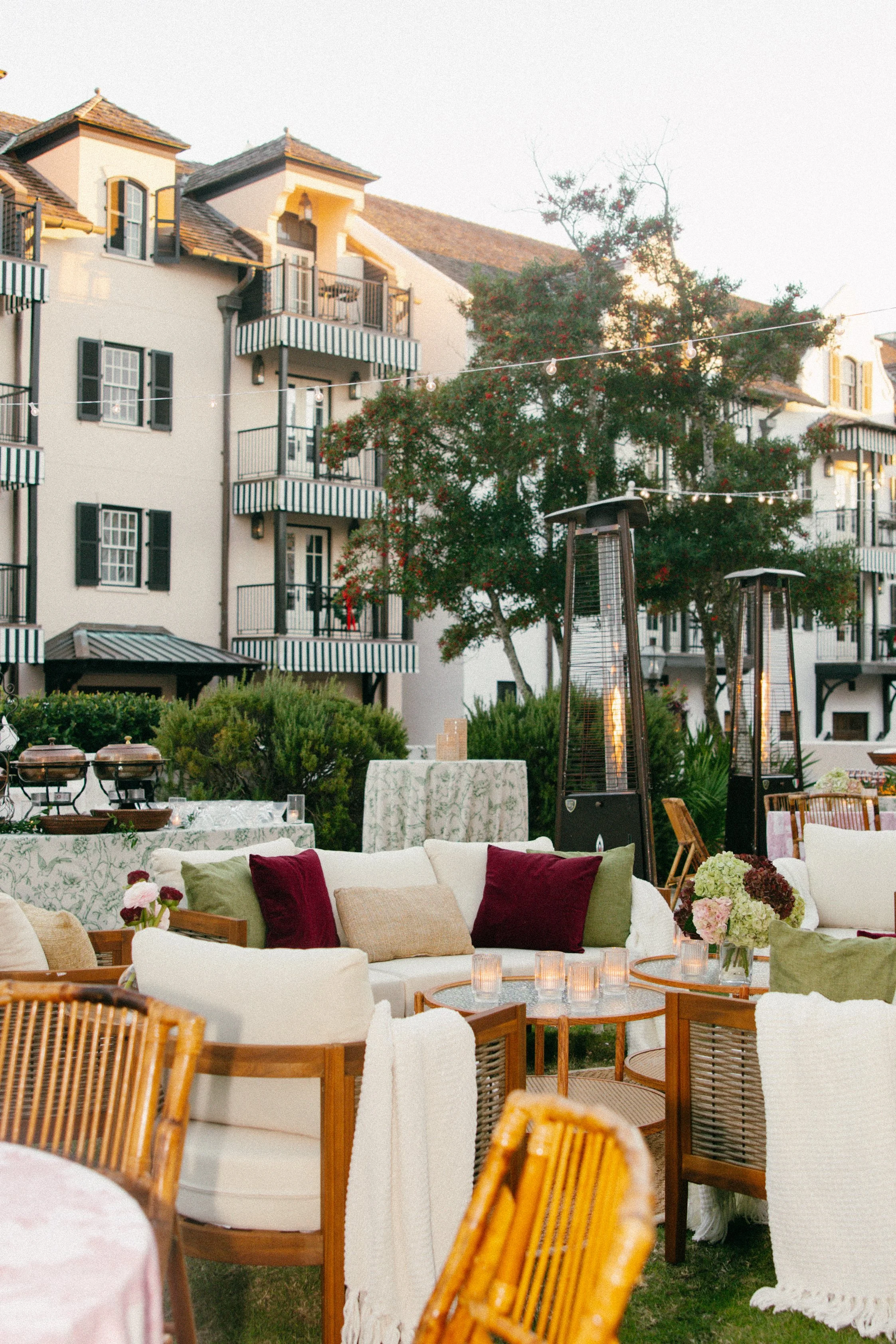 Outdoor event setup with tables, chairs, a sofa with pillows, electric patio heaters, and string lights in front of a residential building with balconies.