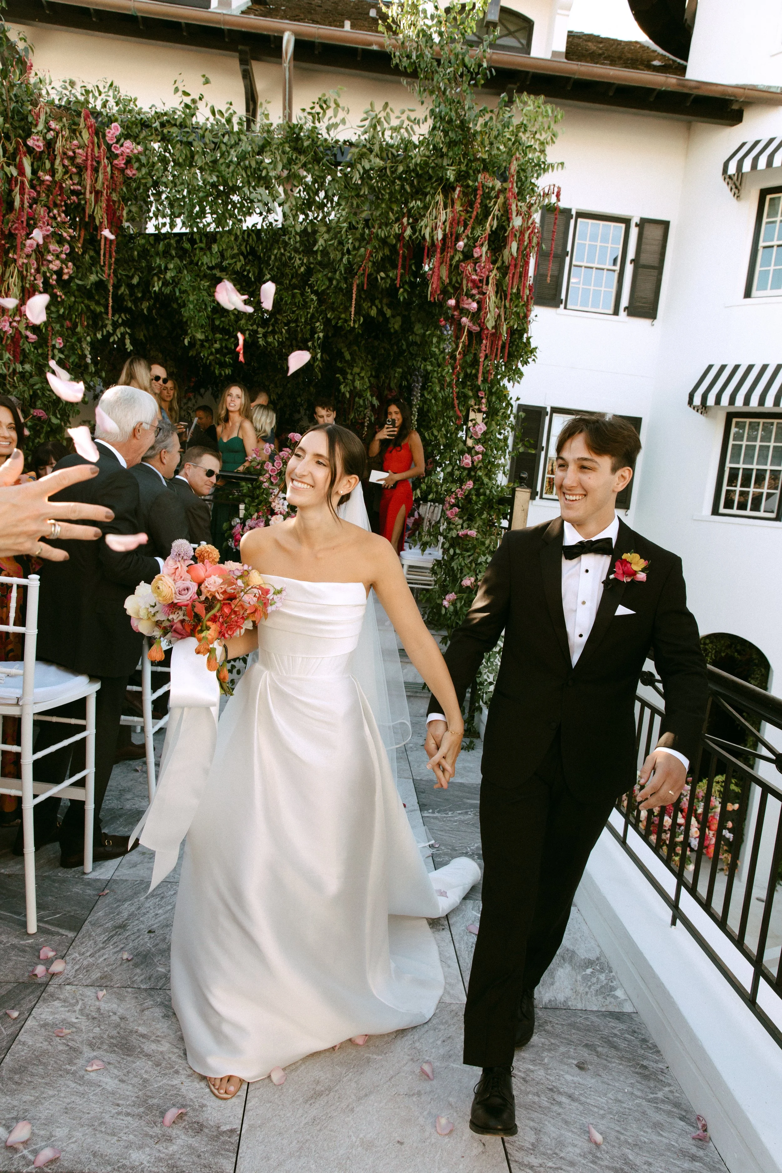 Bride and groom walking down the aisle at their wedding ceremony, holding hands and smiling. The bride is in a white strapless wedding gown holding a bouquet of flowers, and the groom is in a black tuxedo with a bow tie. Guests are seated on either side, some throwing rose petals as they walk.