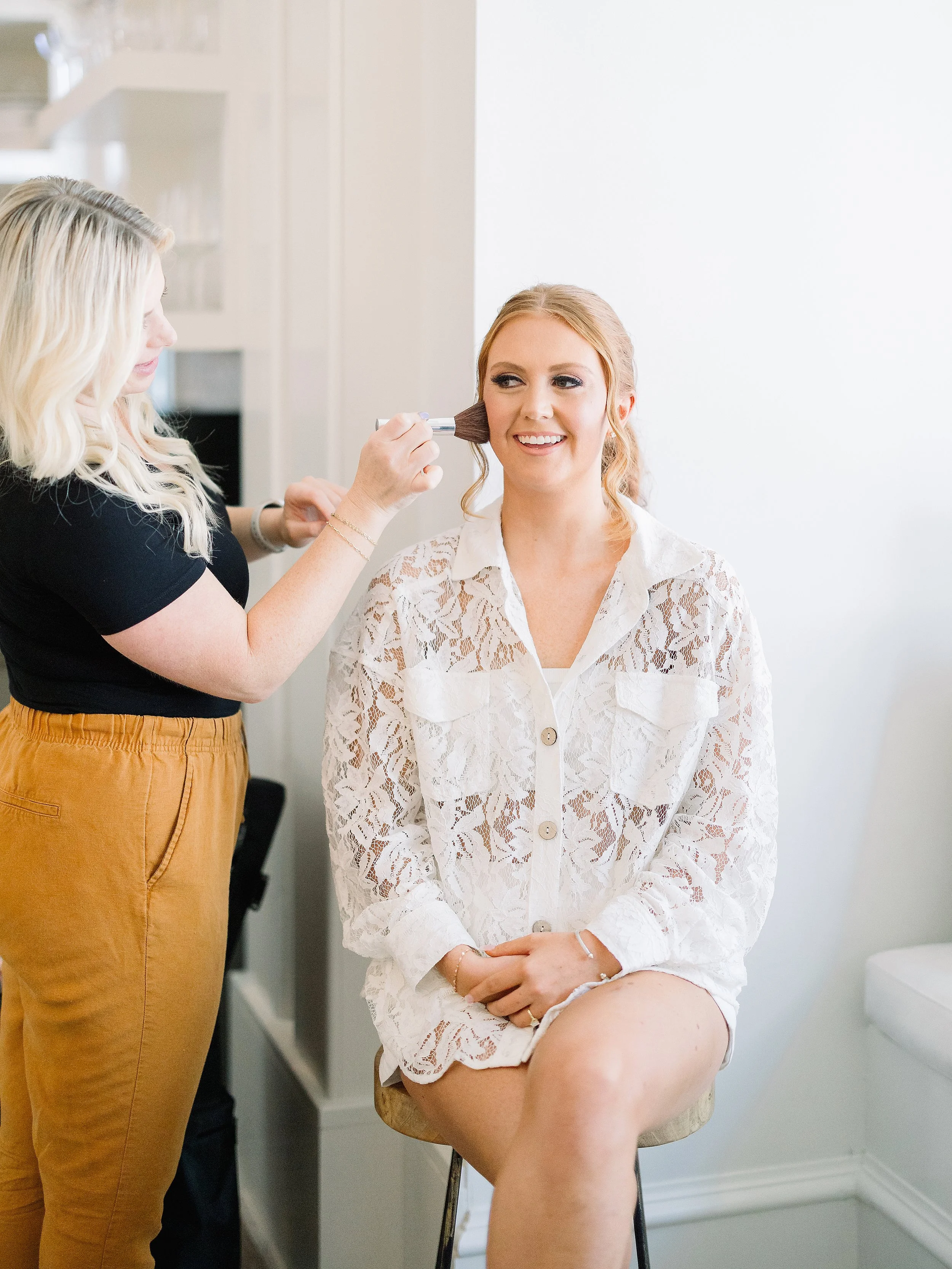 A woman sitting on a stool gets her makeup done by a makeup artist in a bright, white room.