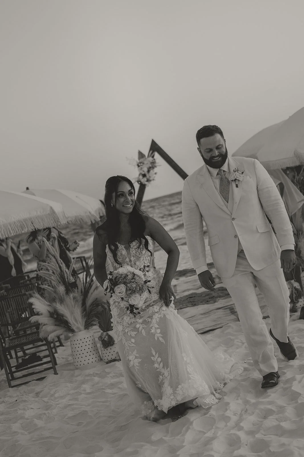 A black and white photo of a bride and groom walking on the beach, holding hands, with umbrellas and beach decor in the background.