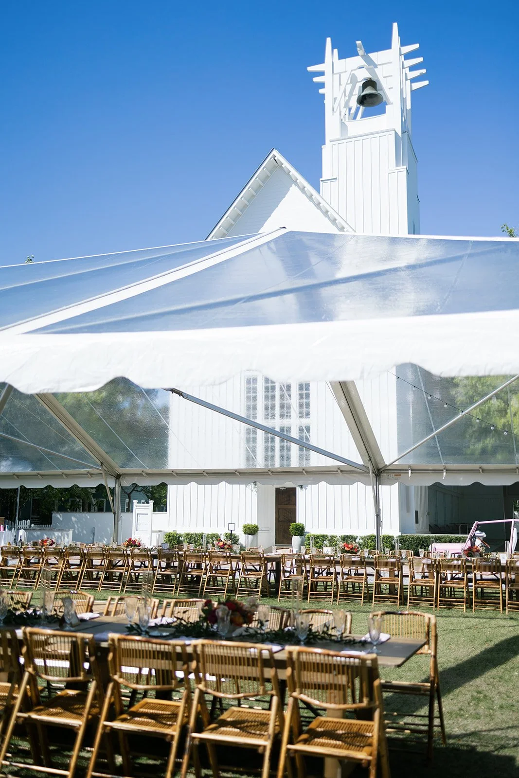 Outdoor wedding setup with wooden chairs and tables under a large white tent, adjacent to a white church building with a bell tower and a clear blue sky.