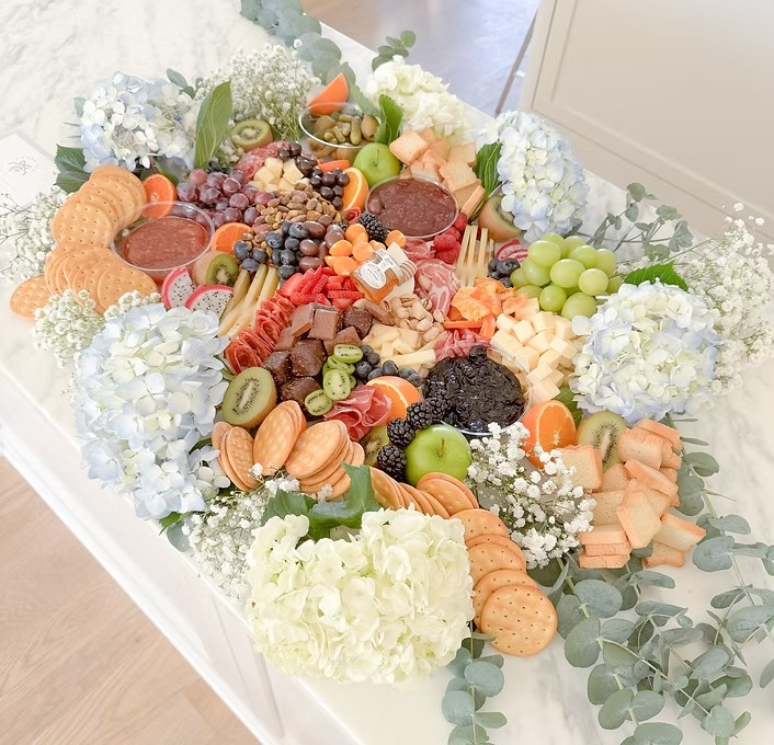 A cheese and fruit platter with crackers, grapes, kiwi, strawberries, and various cheeses, decorated with white and blue hydrangeas and white baby's breath flowers.