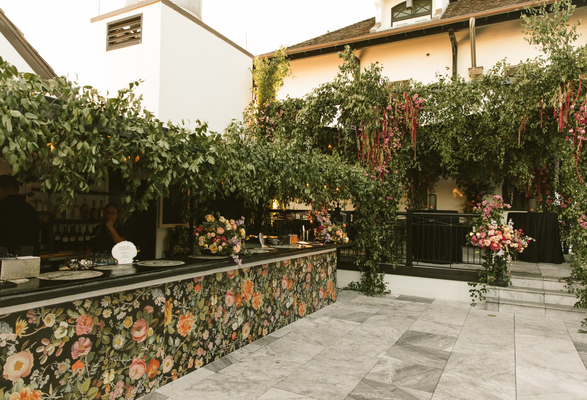 Decorated outdoor venue with floral arrangements, green foliage, and a bar with a floral patterned front, set for a celebration or event.