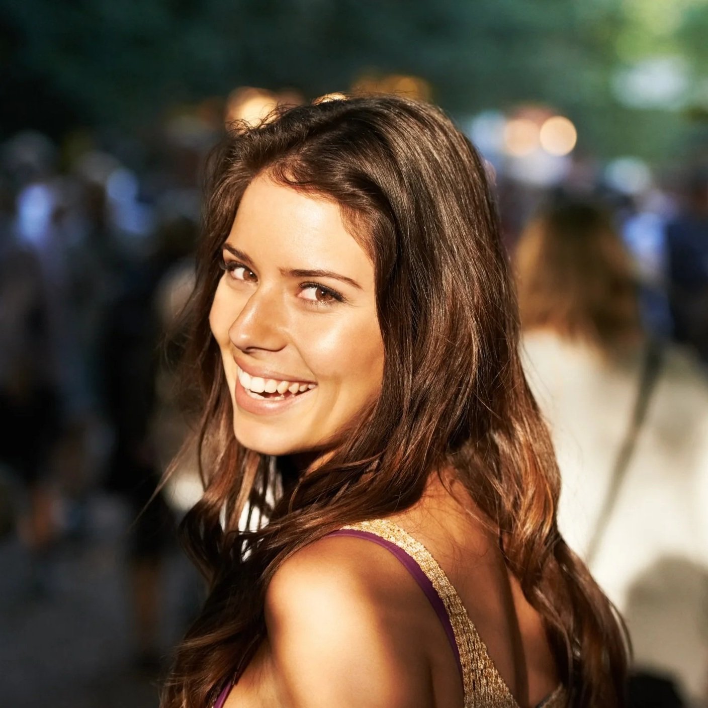 A smiling woman with long, wavy brown hair at an outdoor event during the evening, wearing a sleeveless dress with gold and purple accents, with a blurred crowd in the background.