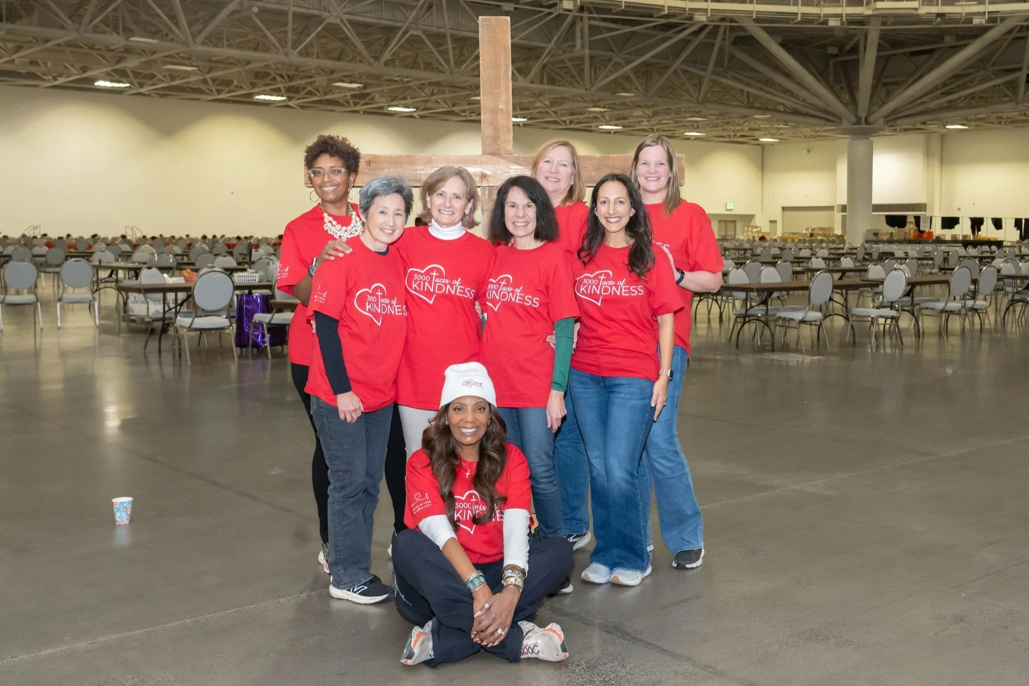 Group of women in red shirts standing together in a large indoor space with empty tables and chairs, holding a wooden cross behind them.