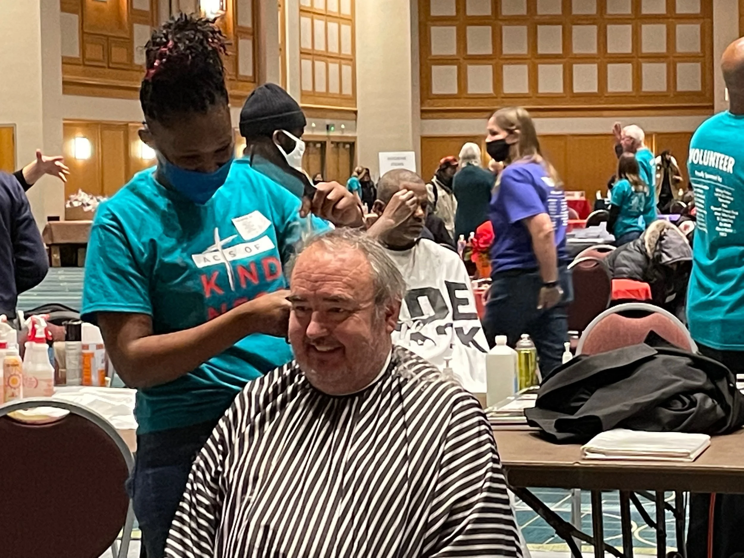 A man receives a haircuts from a woman at a Coated In Love's 3000 Acts of Kindness event, in a large hall filled with people and tables, some of whom are also volunteers.