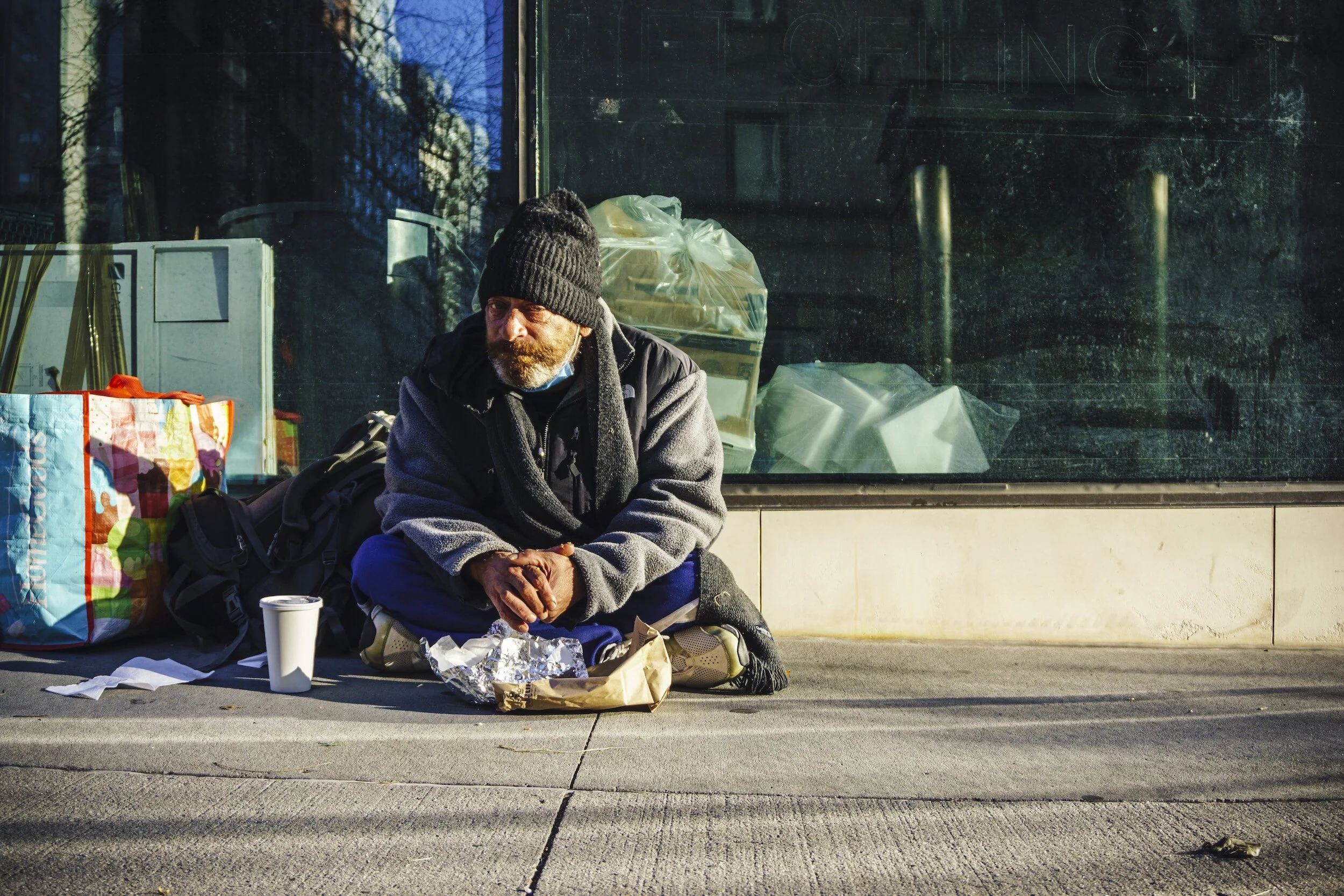A man with a beard wearing a black beanie, gray jacket, and black scarf sits on the sidewalk with food containers, a paper bag, a cup, and a large colorful shopping bag around him, beside a large glass window reflecting nearby trees and buildings.