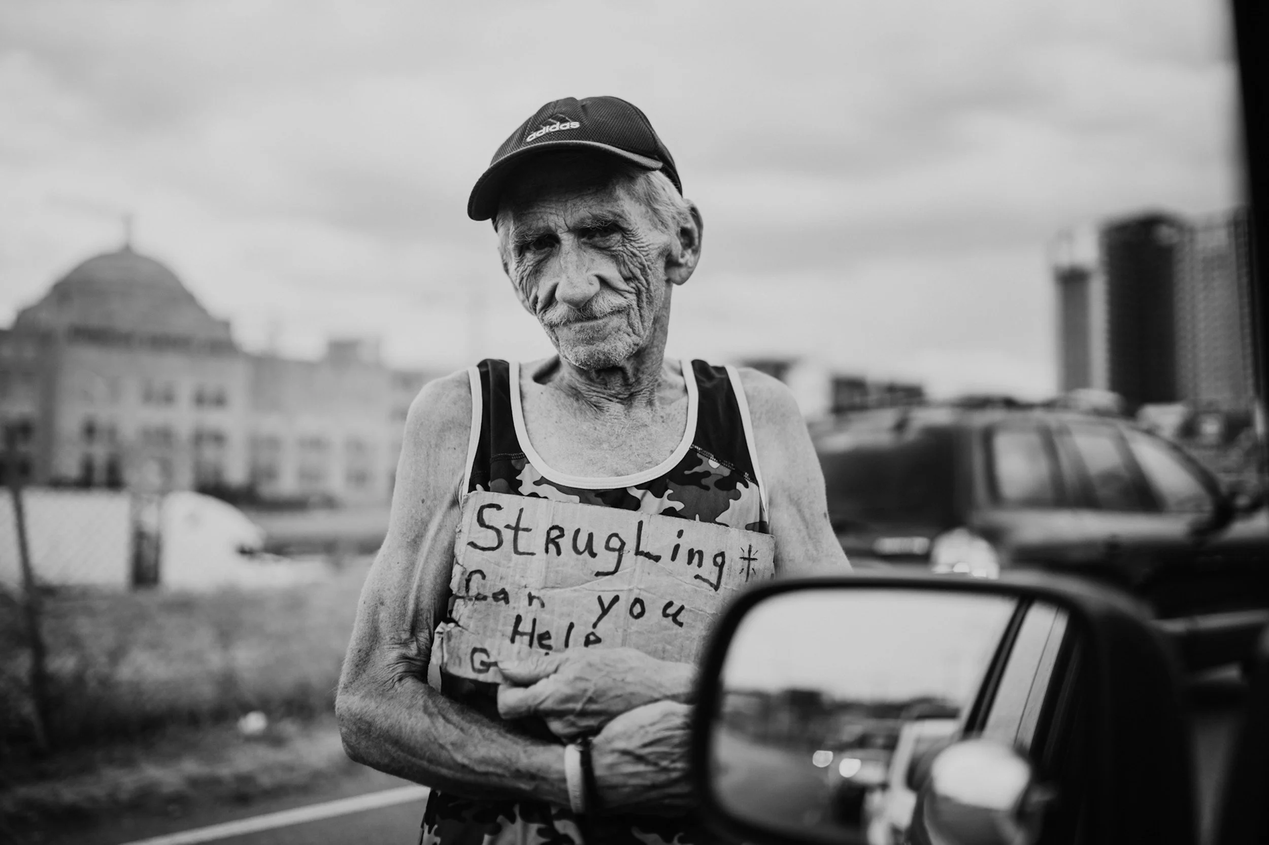 An elderly man with a wrinkled face and wearing a cap and sleeveless shirt holds a handwritten sign that reads, 'Struggling can you help,' while standing outdoors near parked cars and buildings.