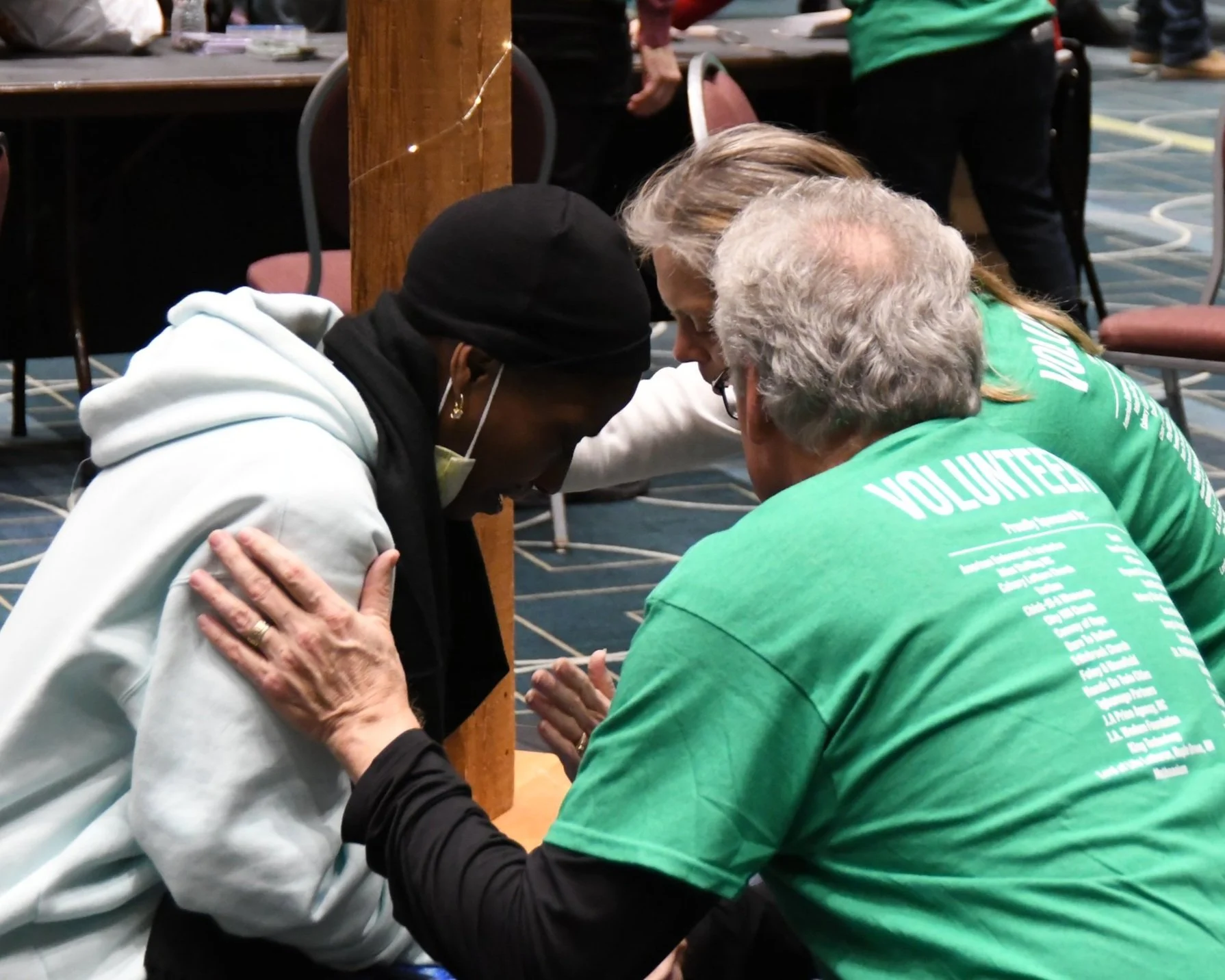 Three volunteers in green shirts kneel around a woman with a black hoodie and face mask, offering comfort or prayer at an indoor event.