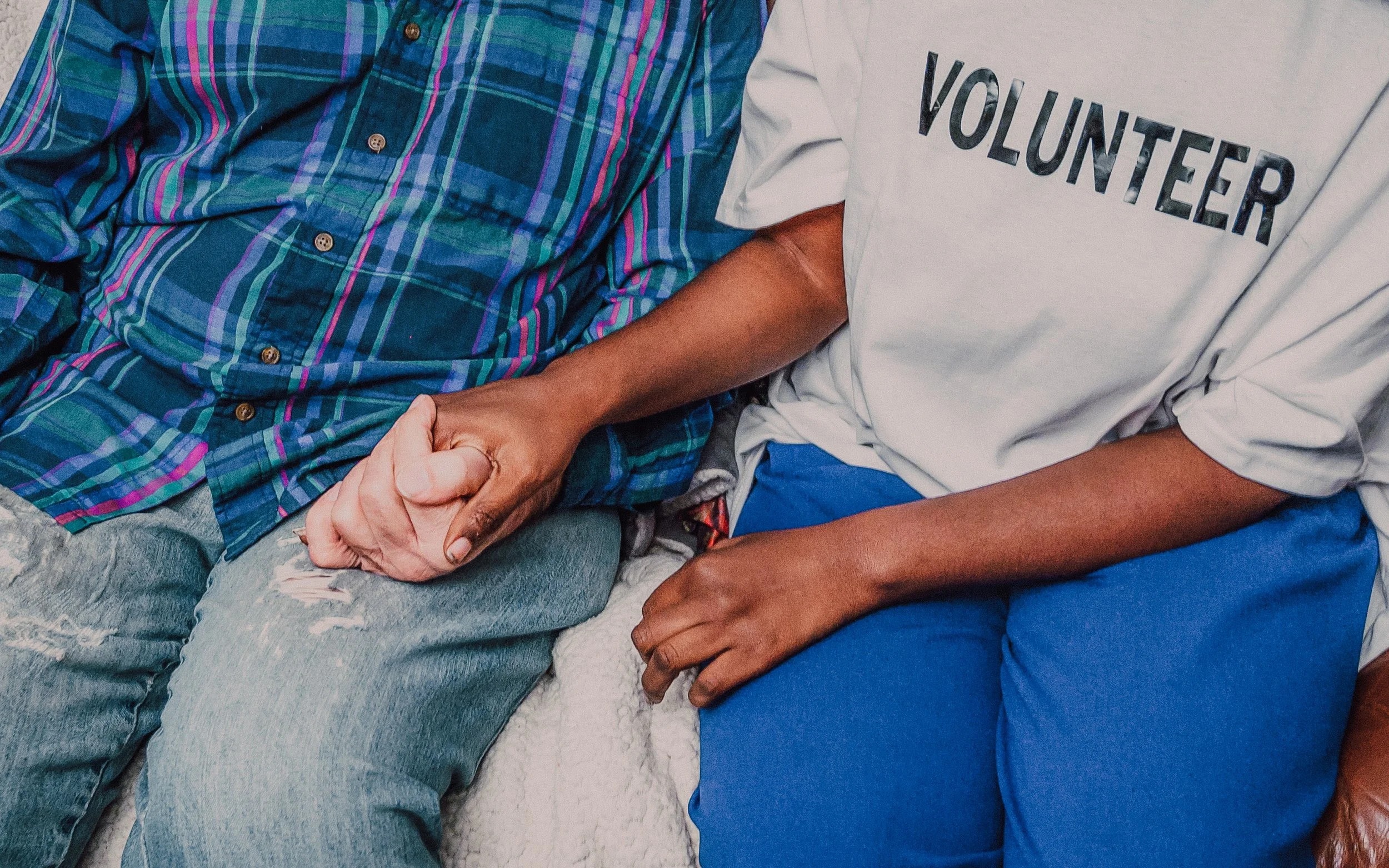 Close-up of two people holding hands, one wearing a plaid shirt and the other a volunteer shirt, sitting close together.