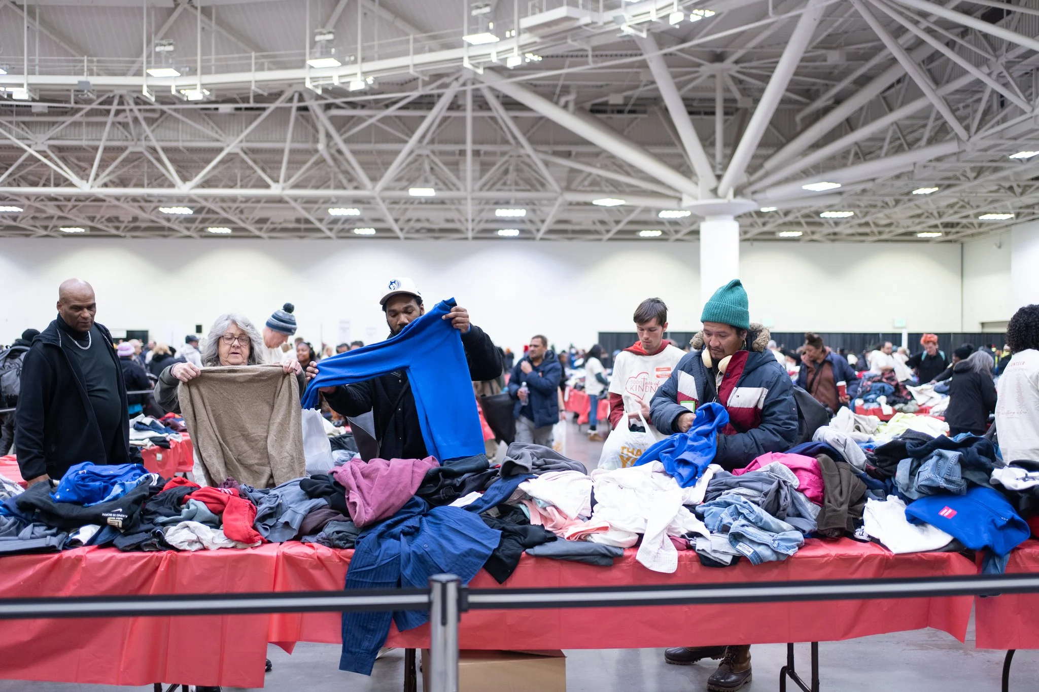 A woman in a green T-shirt talking to a seated woman wearing a gray beanie and black puffer jacket at 3000 Acts of Kindness event with many people and tables in the background.