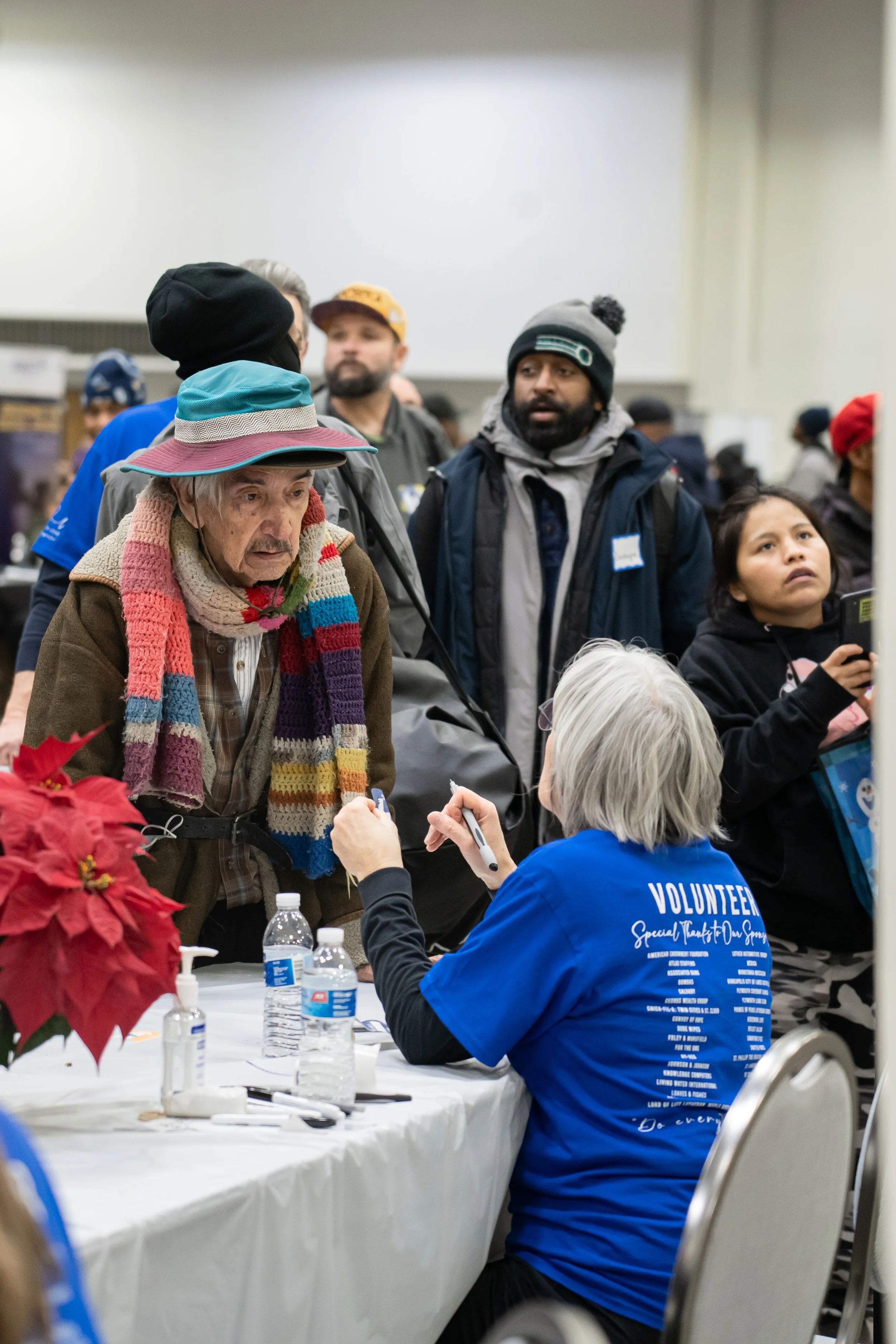 An elderly man wearing a multicolored scarf and a wide-brimmed hat talking to a volunteer sitting at a table with water bottles and pens during a community event, with other people waiting in line behind him.