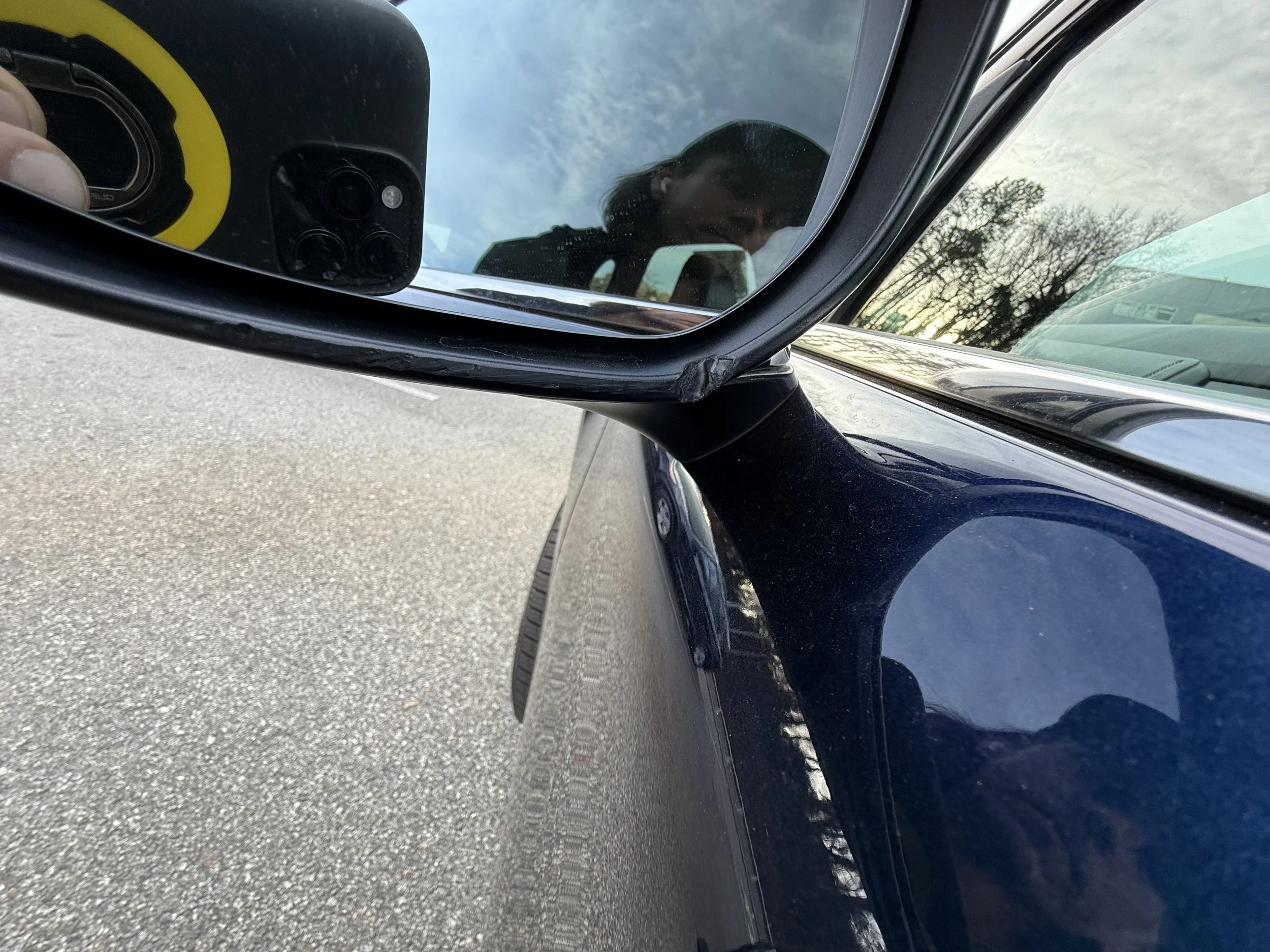 Close-up of a side mirror on a dark-colored vehicle reflecting a person taking the photo, with a person visible in the background.