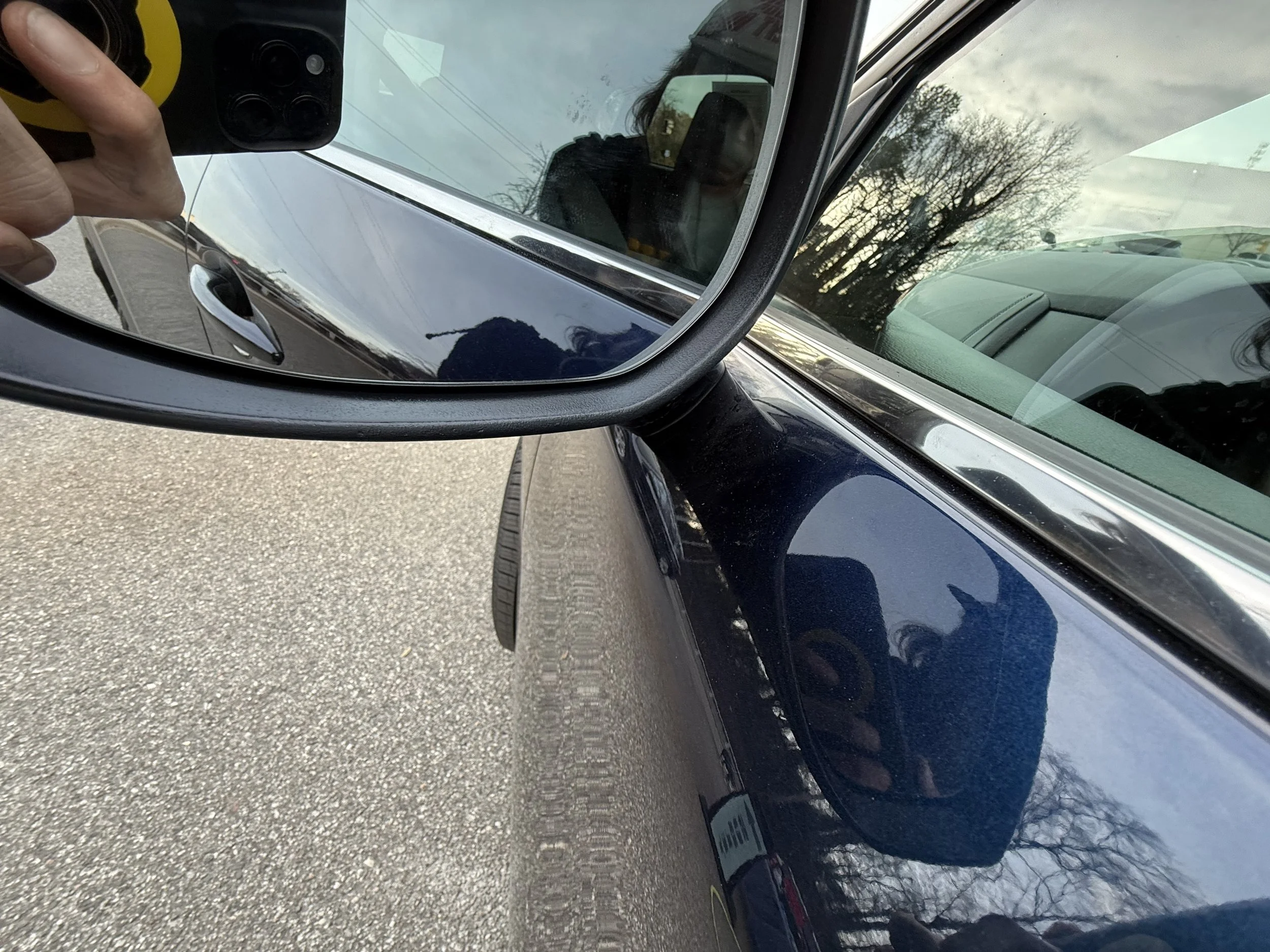 Close-up of a black car's side mirror reflecting the photographer's hand holding a camera, with part of the car's tire and window visible, and trees in the background.