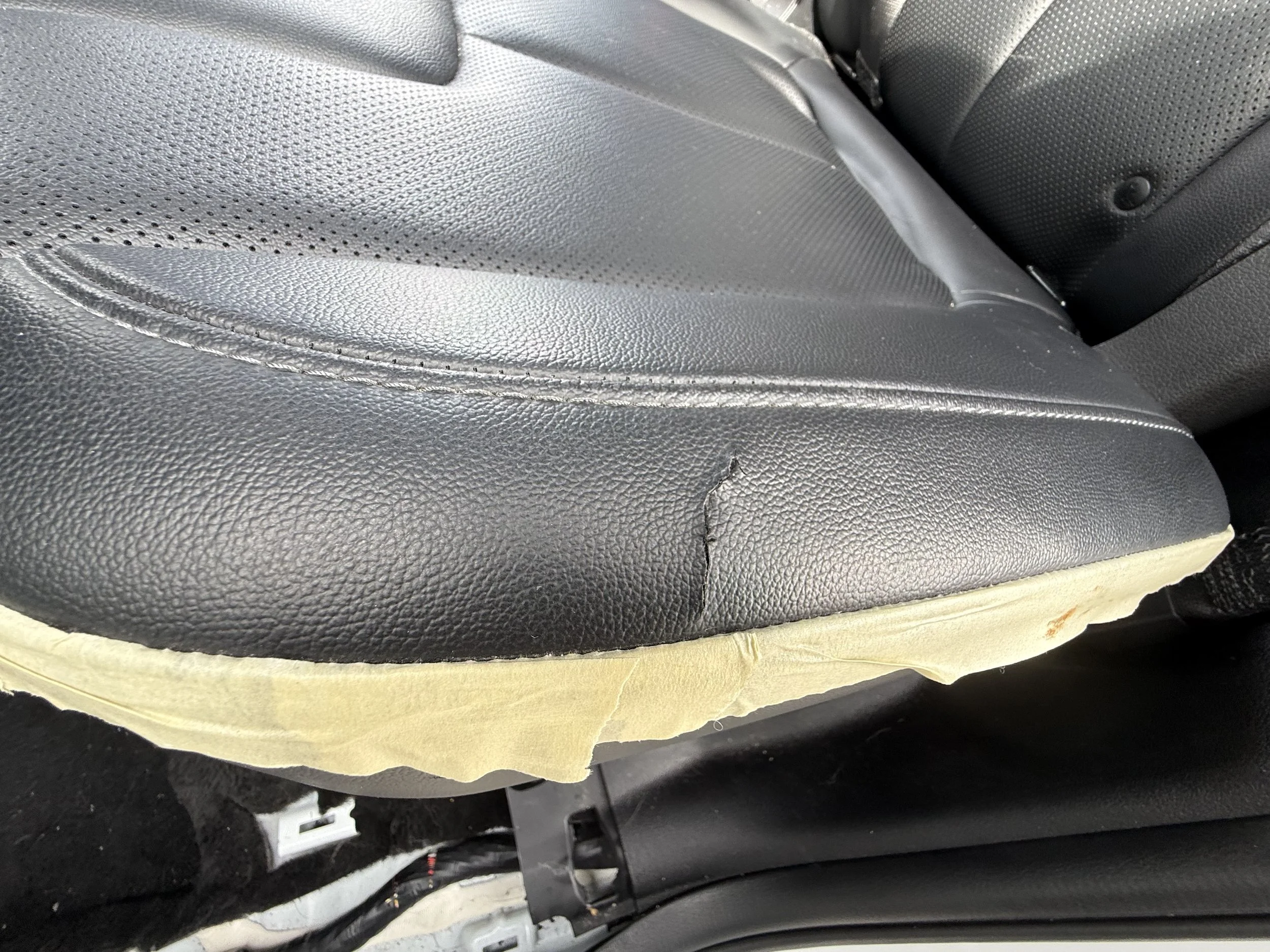 Close-up of a black leather car seat with torn upholstery and masking tape along the edge.