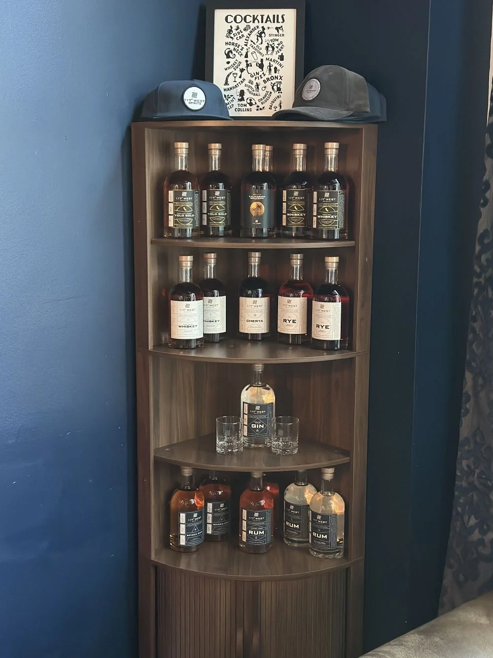 A wooden liquor shelf with bottles of whiskey, gin, and rum, two caps on top, and a framed cocktail poster behind.