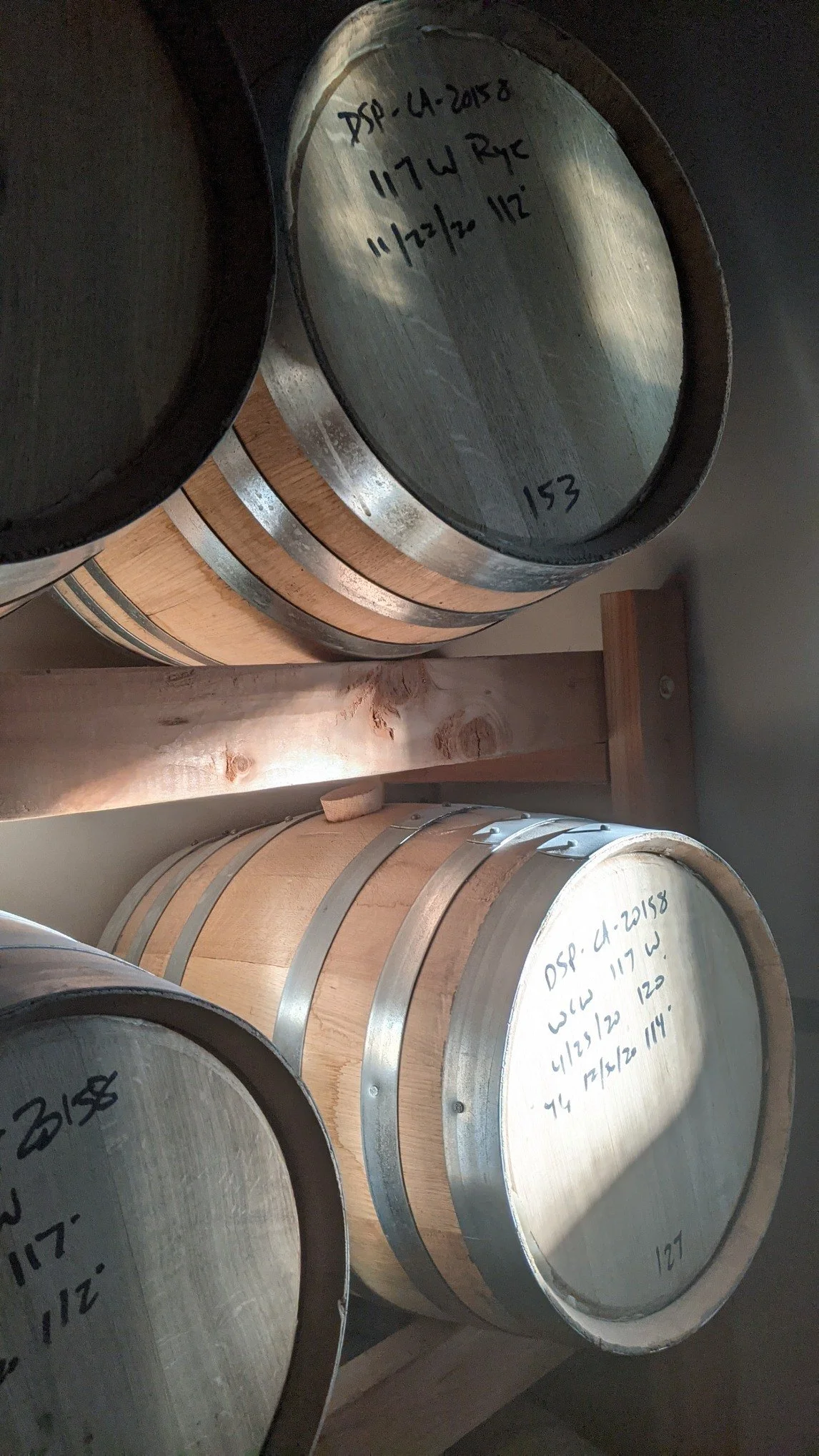 Wooden barrels stacked in a storage room, each marked with handwritten notes, possibly indicating whiskey or spirit types.