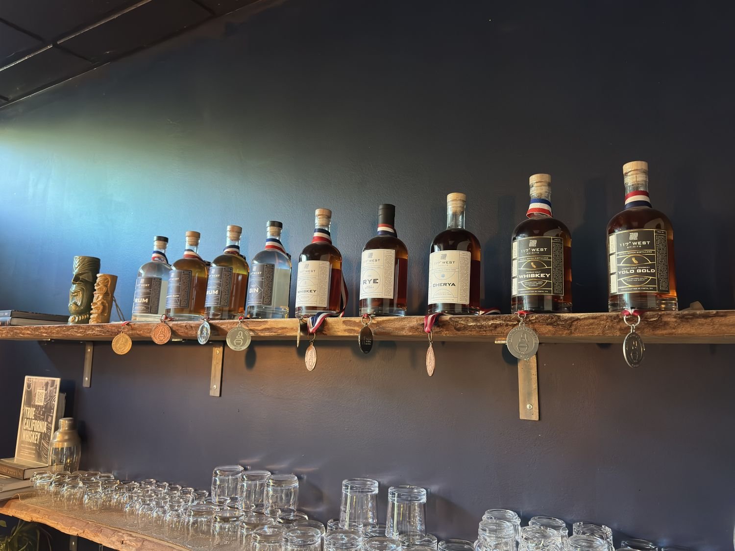 Display of whiskey bottles on a wooden shelf above a row of empty glasses in a bar setting.