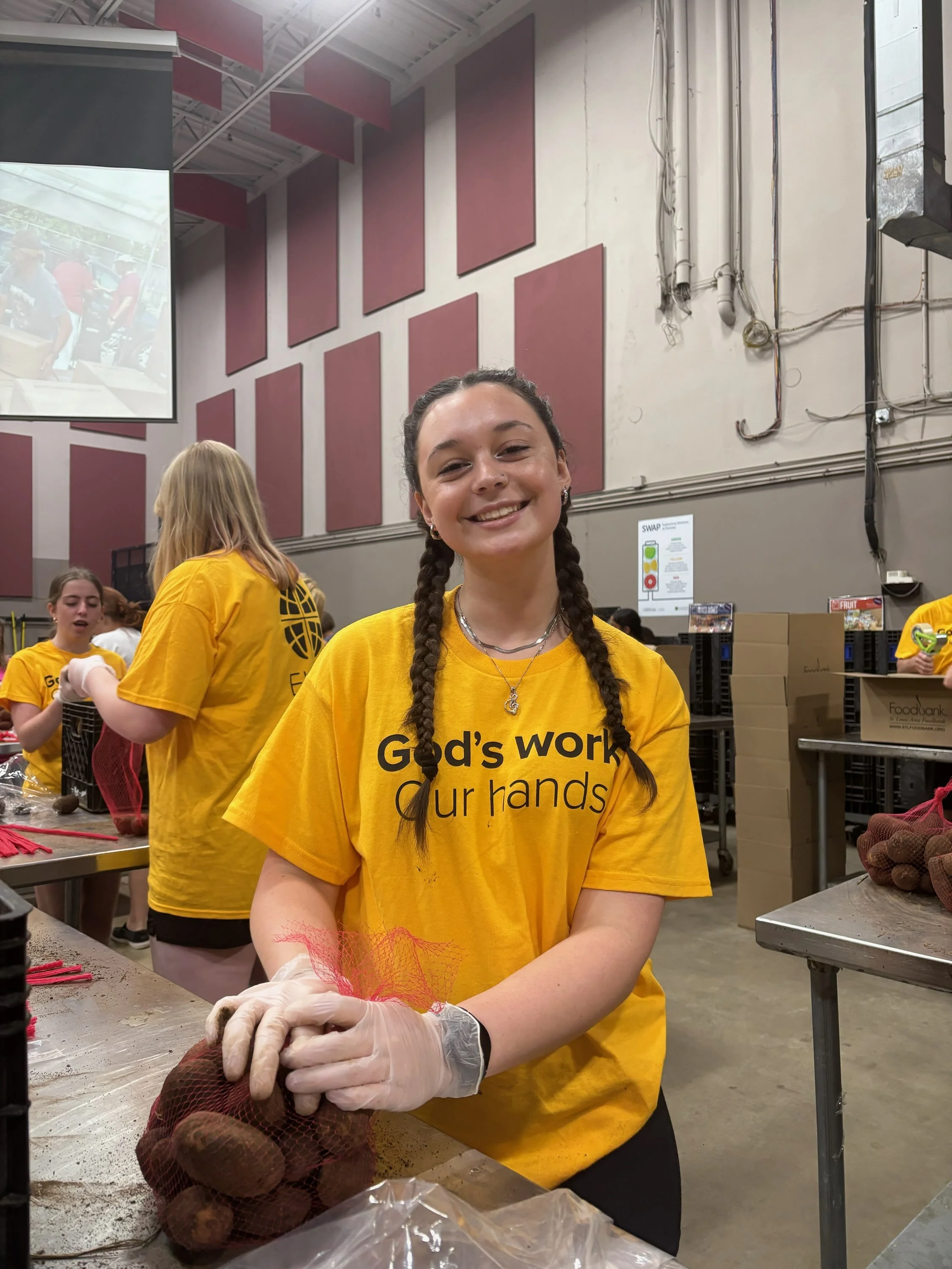 A young woman with braided hair smiling while bagging potatoes at a volunteering event, wearing a yellow shirt with the text 'God's work, our hands.'