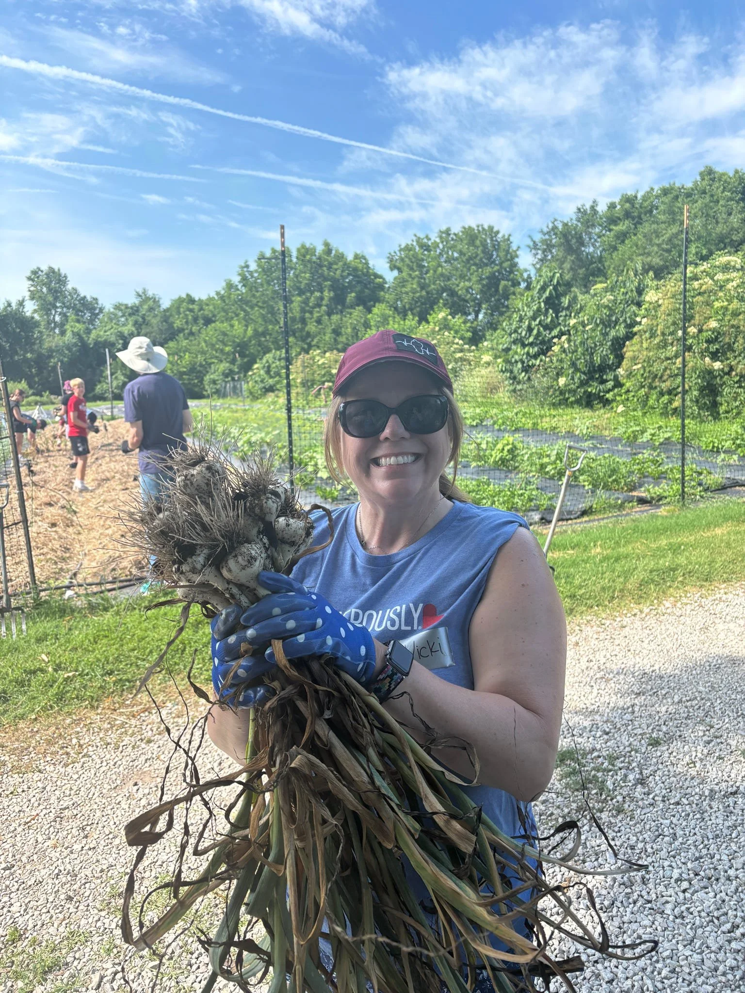 Woman in sunglasses, wearing a blue sleeveless shirt, holding a bunch of garlic bulbs, smiling outdoors in a garden. There are other people working in the background.