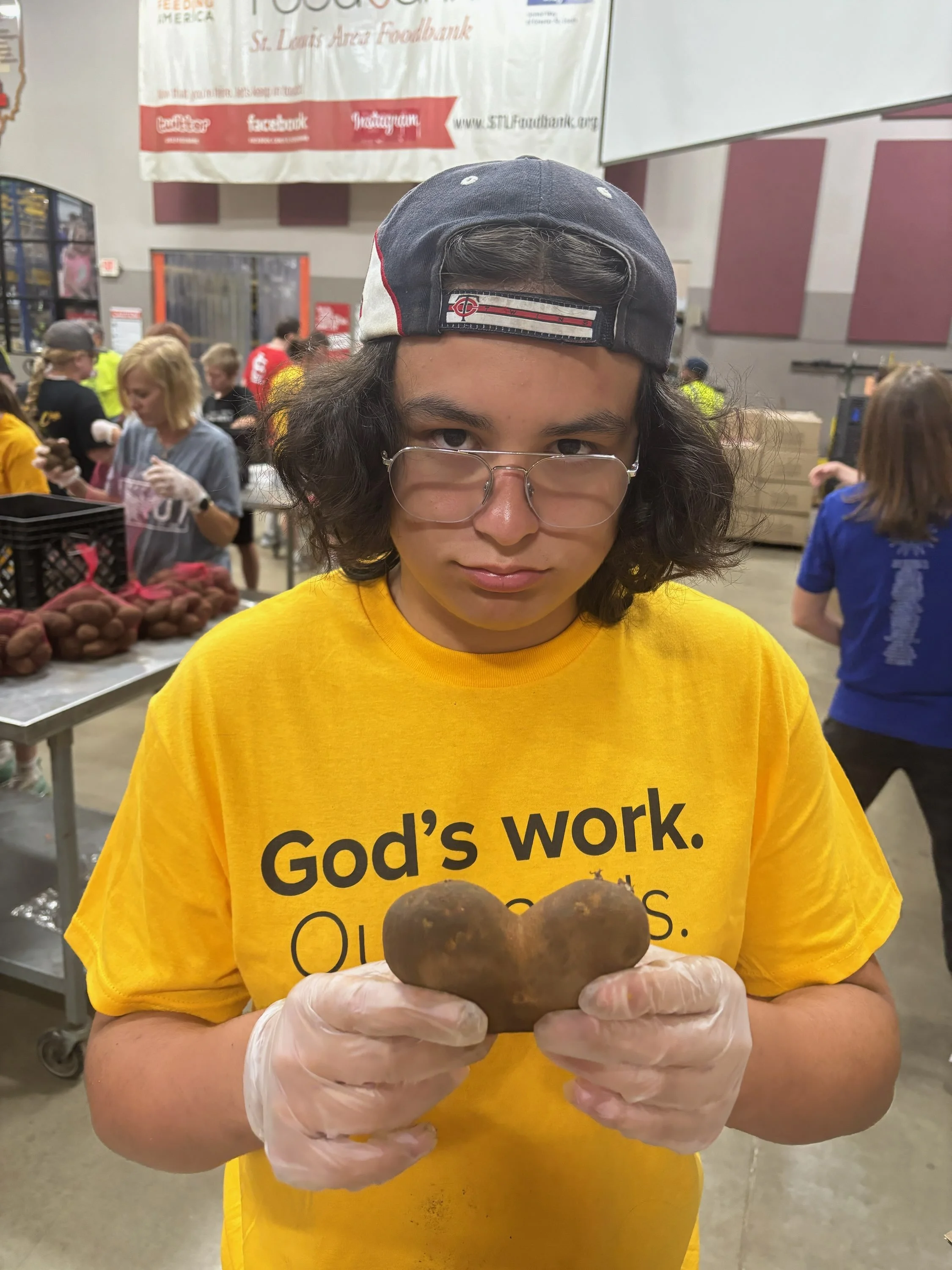 A young person with shoulder-length dark hair, wearing glasses, a backwards navy baseball cap, and a yellow T-shirt with the text "God's work. Our hands." is holding two large potatoes shaped like hearts in a food bank or community center, with other volunteers working in the background.
