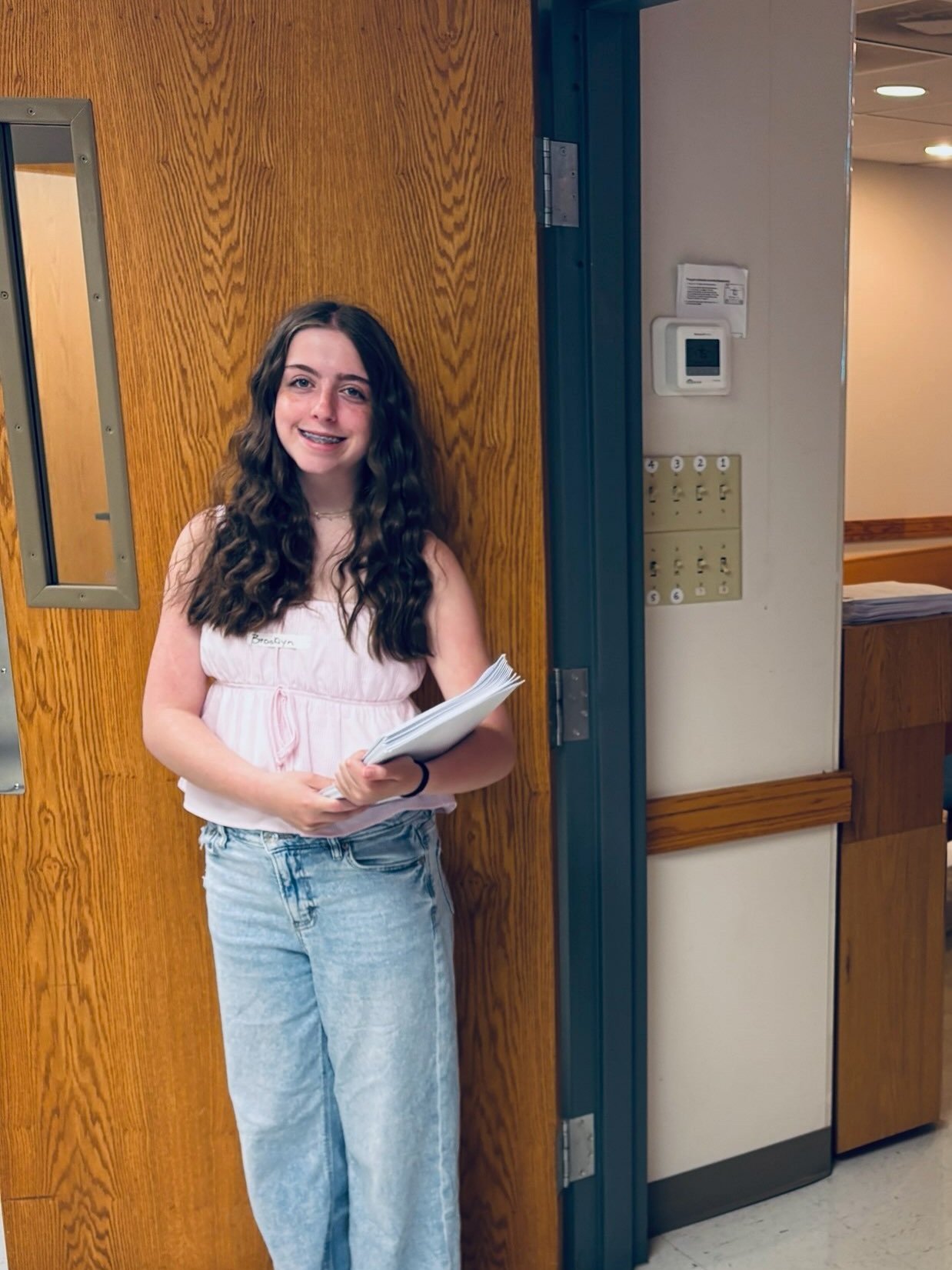 A young girl with long curly brown hair, wearing a pink sleeveless top and light blue jeans, standing in front of a wooden door, holding a stack of white papers and smiling at the camera.