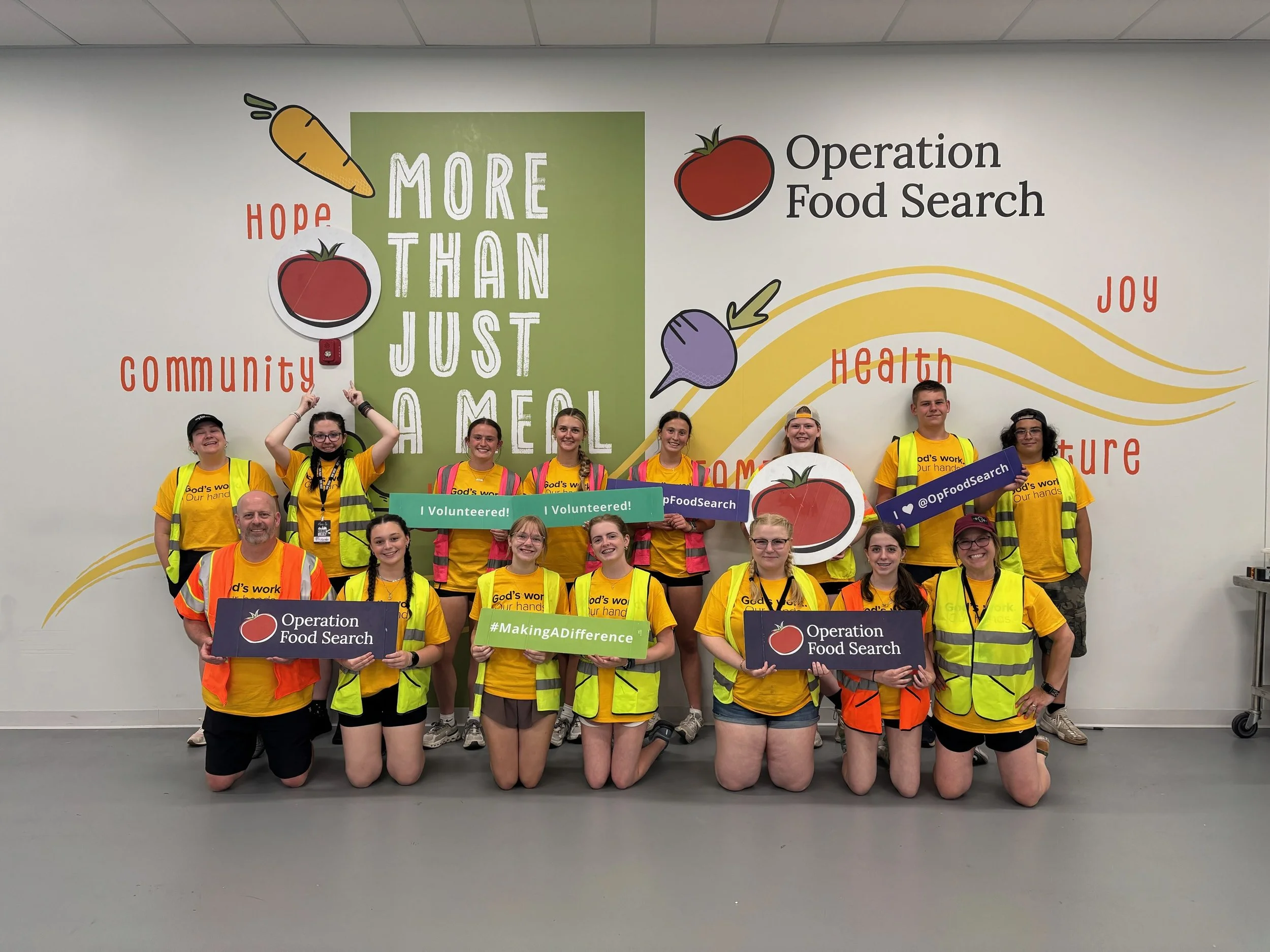 Group of volunteers wearing yellow shirts and safety vests, holding signs for Operation Food Search, in front of a wall with colorful food-themed artwork and motivational phrases.