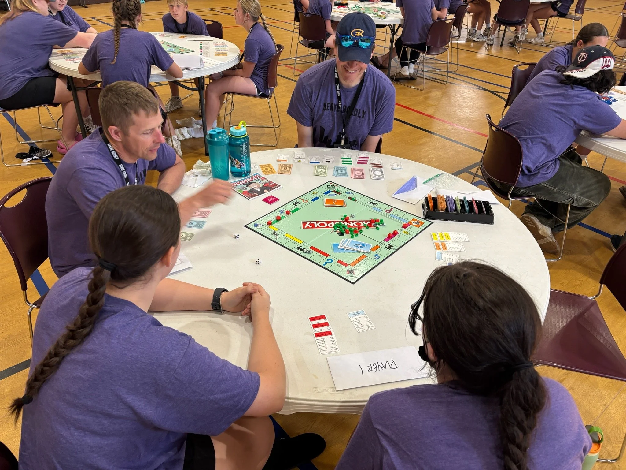 People gathered around a round table playing Monopoly in a gymnasium, with additional groups playing nearby. The group includes a woman with a braid, a man with glasses, and a woman in a purple shirt. The table is set with Monopoly game pieces, money, and game cards.