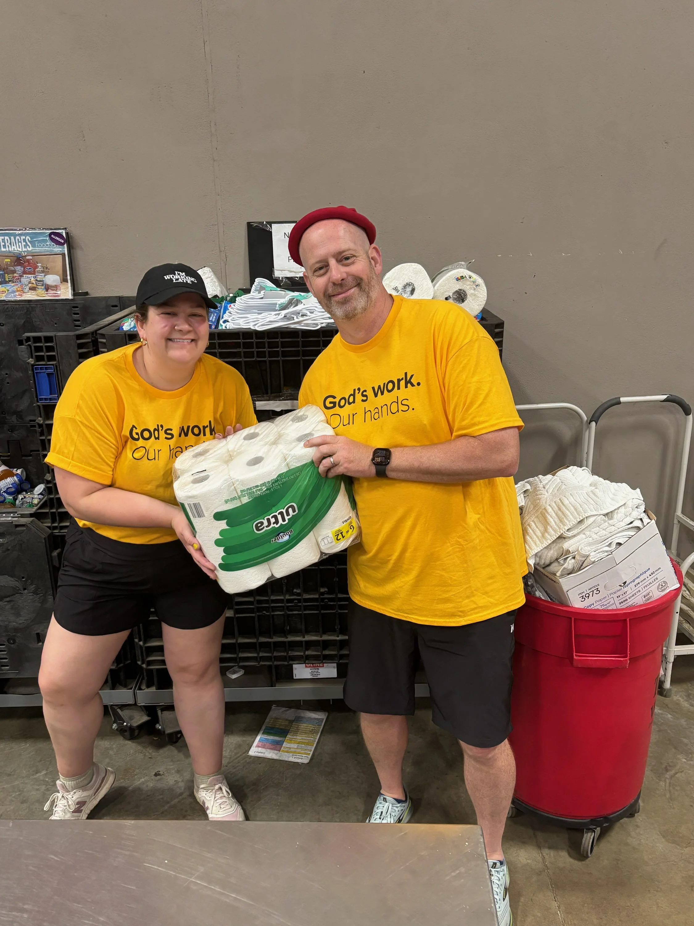 Two volunteers in yellow shirts volunteering at a food bank, holding a pack of paper towels together. The man is wearing a red hat and the woman is wearing a black cap. They are standing in front of shelves filled with supplies and a large red bin.